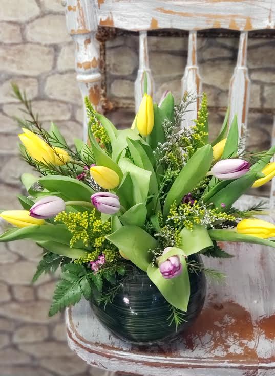 Low arrangement of yellow and purple-tipped tulips in a round glass vase on a rustic white chair