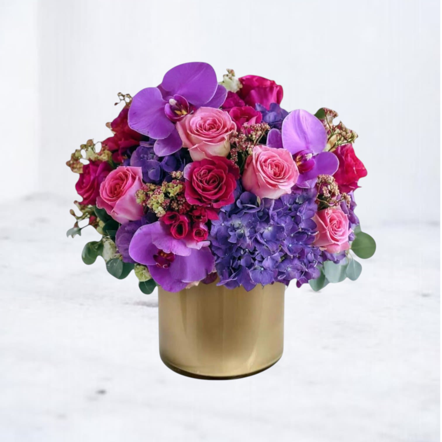 Royal Amethyst - A jewel-toned cluster of purple phalaenopsis orchids, vivid purple hydrangeas, and pink and red roses creates an immediate sense of luxury-perfect as an anniversary arrangement or a romantic birthday surprise. This flower arrangement for love and romance also reads as a confident congratulatory gift or a cheerful retirement centerpiece, the kind that makes the person you're thinking of feel truly seen.  Dense, rounded and layered, the composition pairs large orchid blooms with lush hydrangea heads and mixed roses and spray roses for texture and depth; small accent flowers and subtle foliage peek through without competing with the main blooms. Housed in a smooth gold metallic cylinder vase, the design measures as a compact yet impactful display-modern, sculptural, and ready to sit on a dining table or office reception. Order for same-day delivery where available to celebrate birthdays, anniversaries, retirements, or any special moment that calls for a striking, elegant statement.