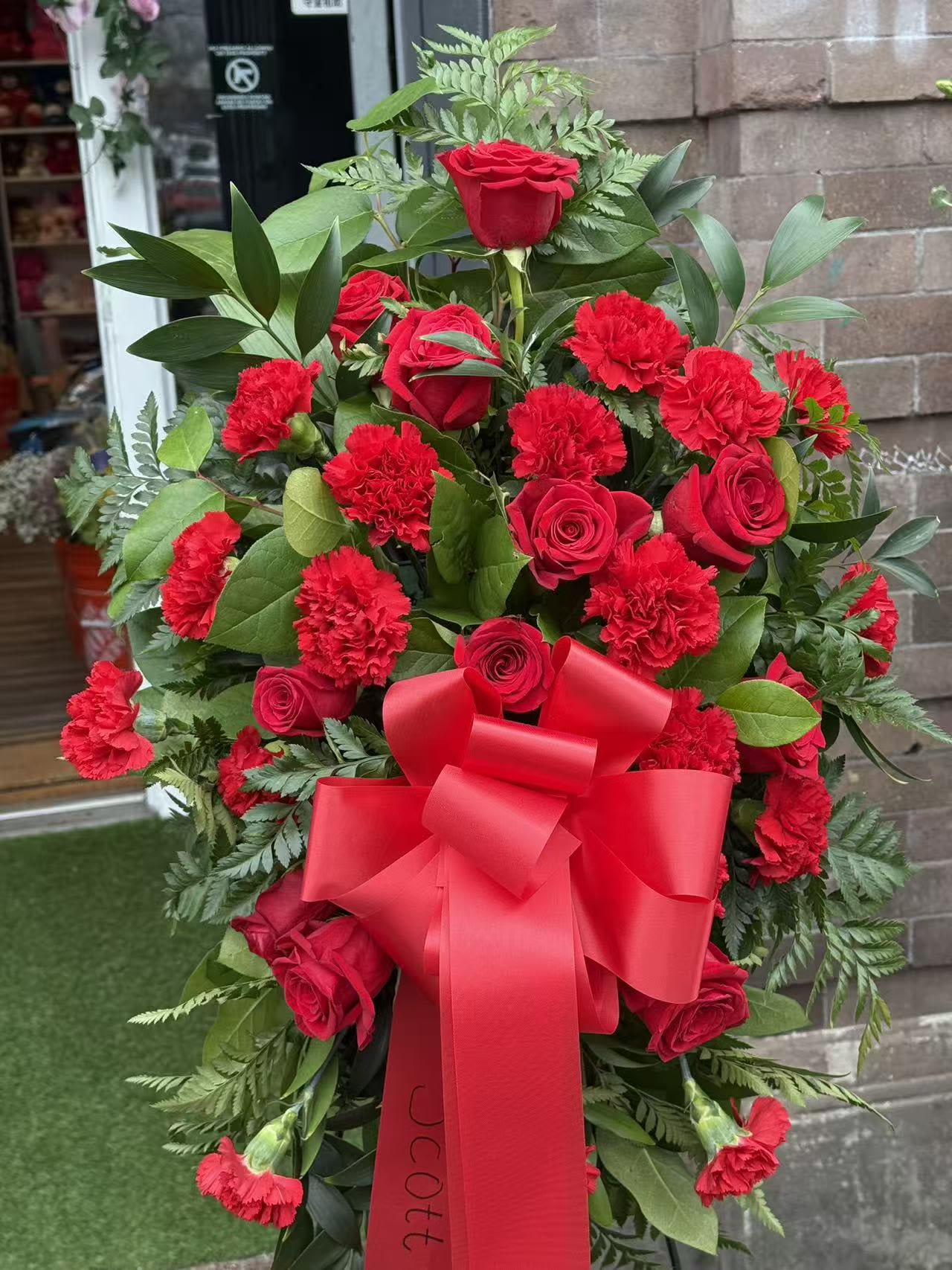 Eternal Love Tribute Spray - A timeless and elegant standing spray featuring premium red roses and vibrant red carnations, beautifully arranged with lush seasonal greenery and finished with a classic red ribbon. This arrangement expresses deep love, respect, and remembrance, making it a meaningful tribute for funeral services and memorials.