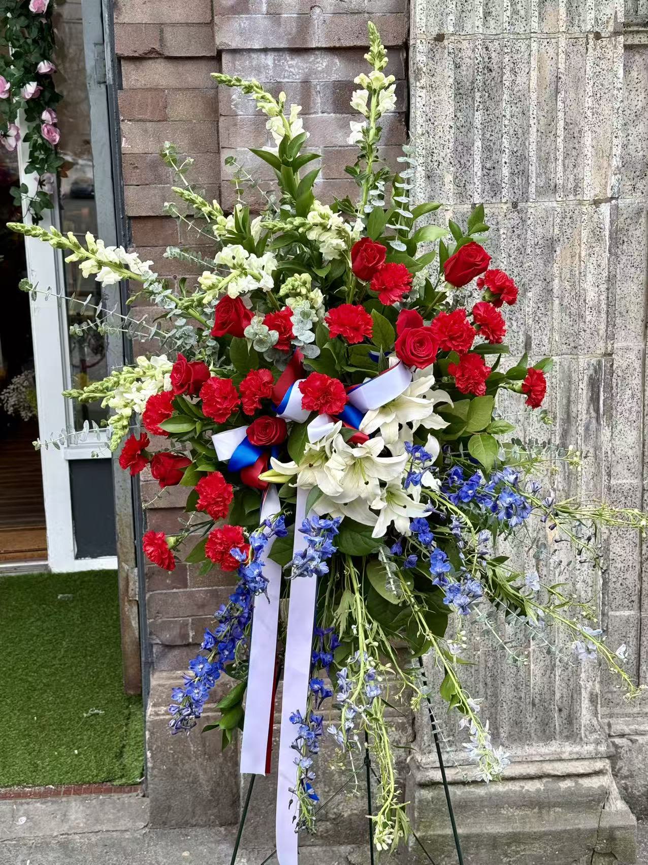 Patriotic Honor Standing Spray - A striking and meaningful standing spray designed in a classic red, white, and blue palette. Featuring premium red roses, red carnations, elegant white lilies, and seasonal blue delphinium, accented with lush greenery and a patriotic ribbon. This arrangement honors a life of dignity, service, and love—perfect for memorials and funeral services.