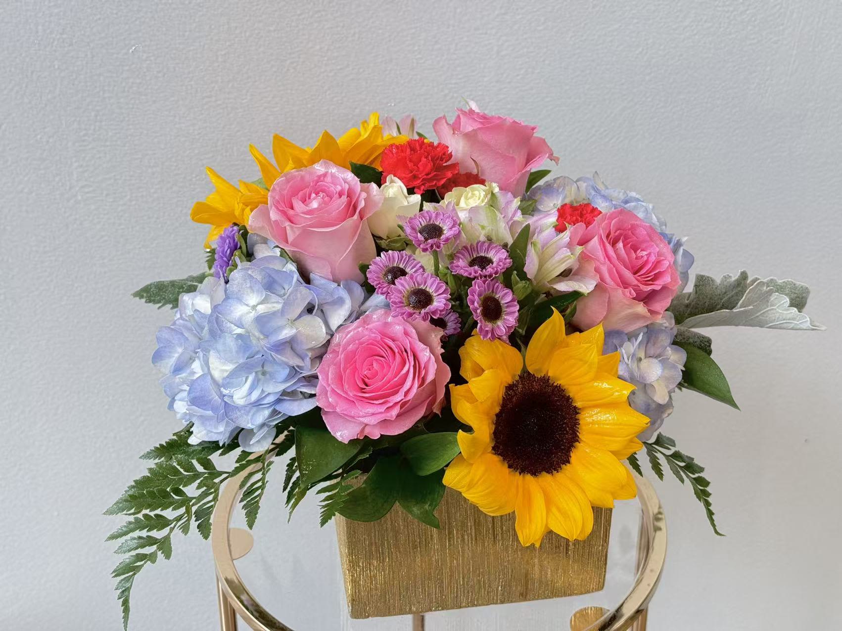 Sweet Sunshine Floral Box - A bright and joyful floral arrangement featuring pink roses, blue hydrangea, sunflowers, and seasonal blooms, beautifully designed in a modern gold container. This cheerful design blends softness and sunshine, making it perfect for birthdays, celebrations, or sending a warm and thoughtful surprise.