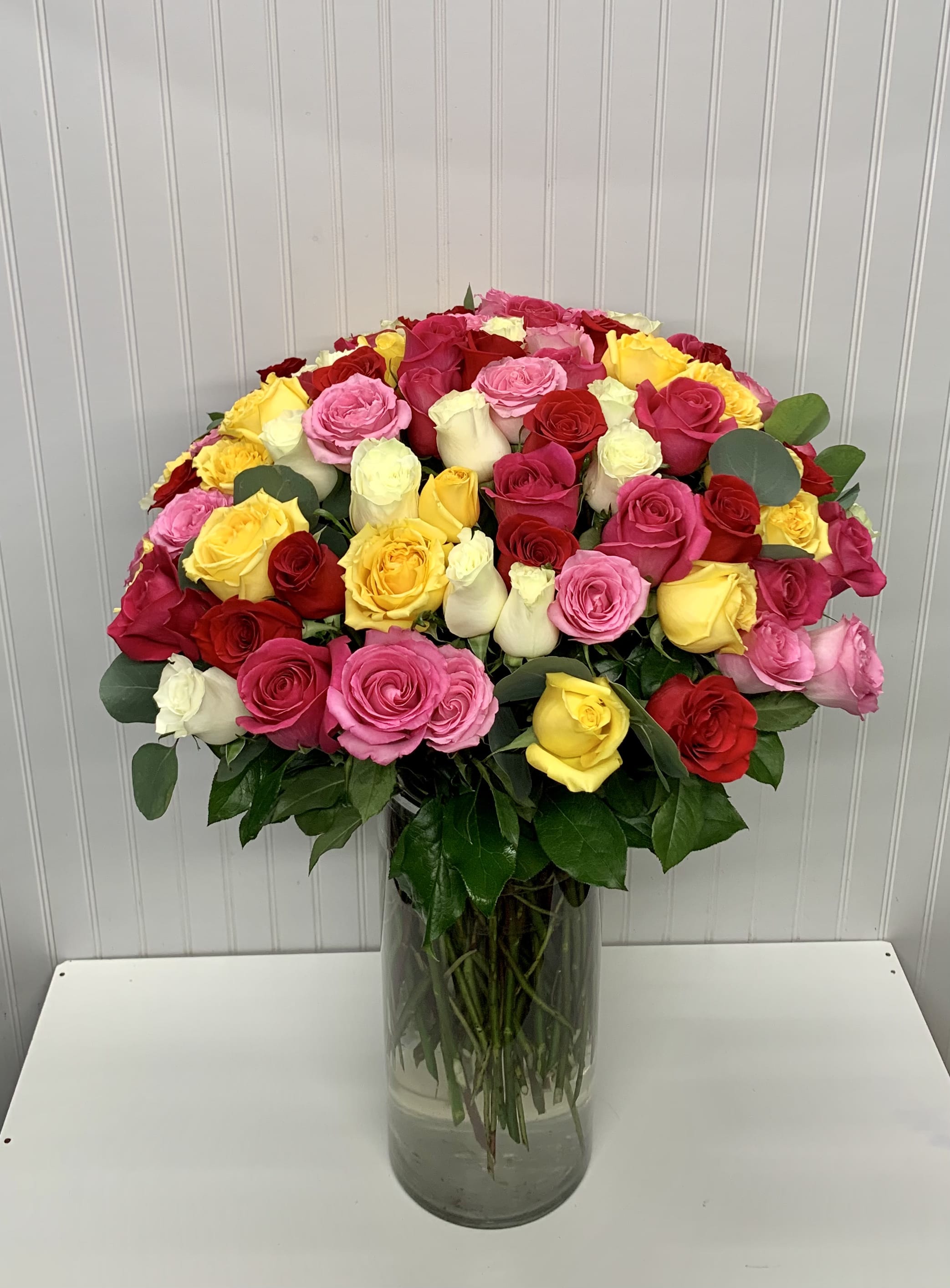 Tall clear vase filled with a large, dome-shaped arrangement of red, pink, yellow and white roses.