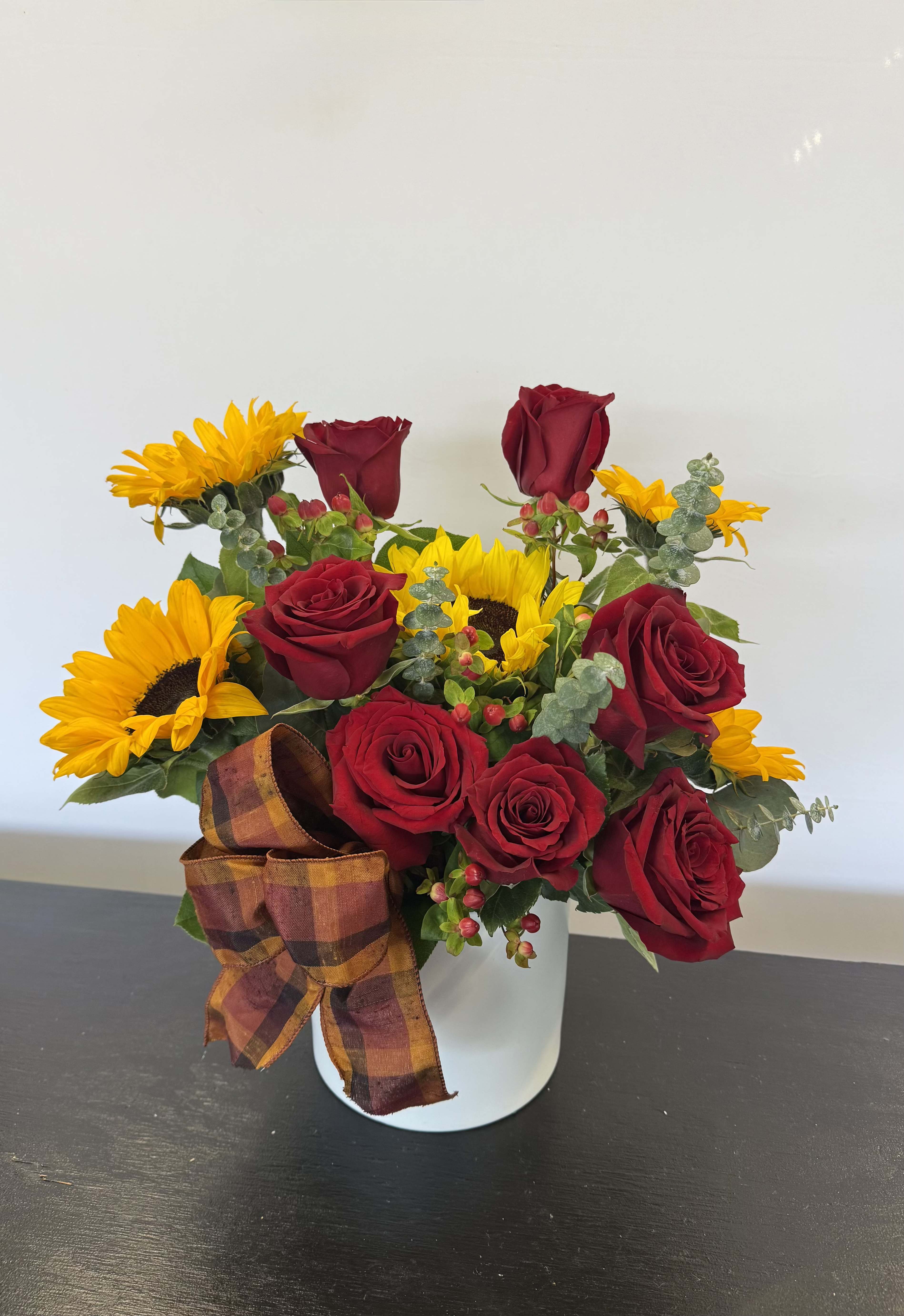 Arrangement of red roses and yellow sunflowers in a white container with a plaid bow.