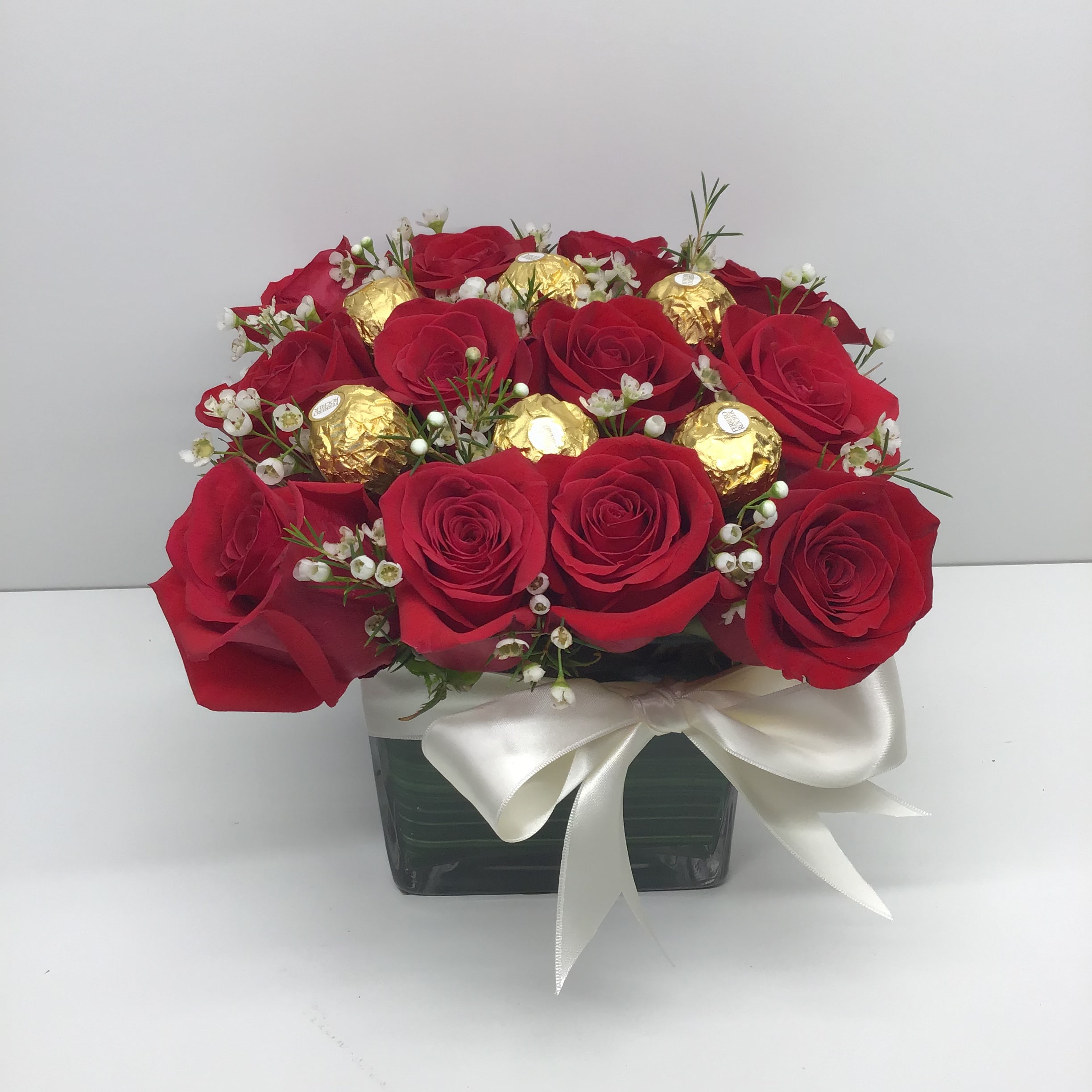 Compact arrangement of red roses and gold-wrapped chocolates in a square vase with a white bow
