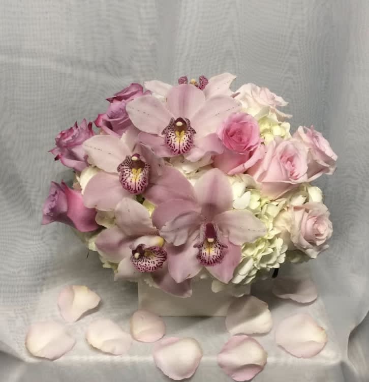 Low arrangement of pink orchids, roses, and white hydrangeas in a white cube vase with scattered petals.