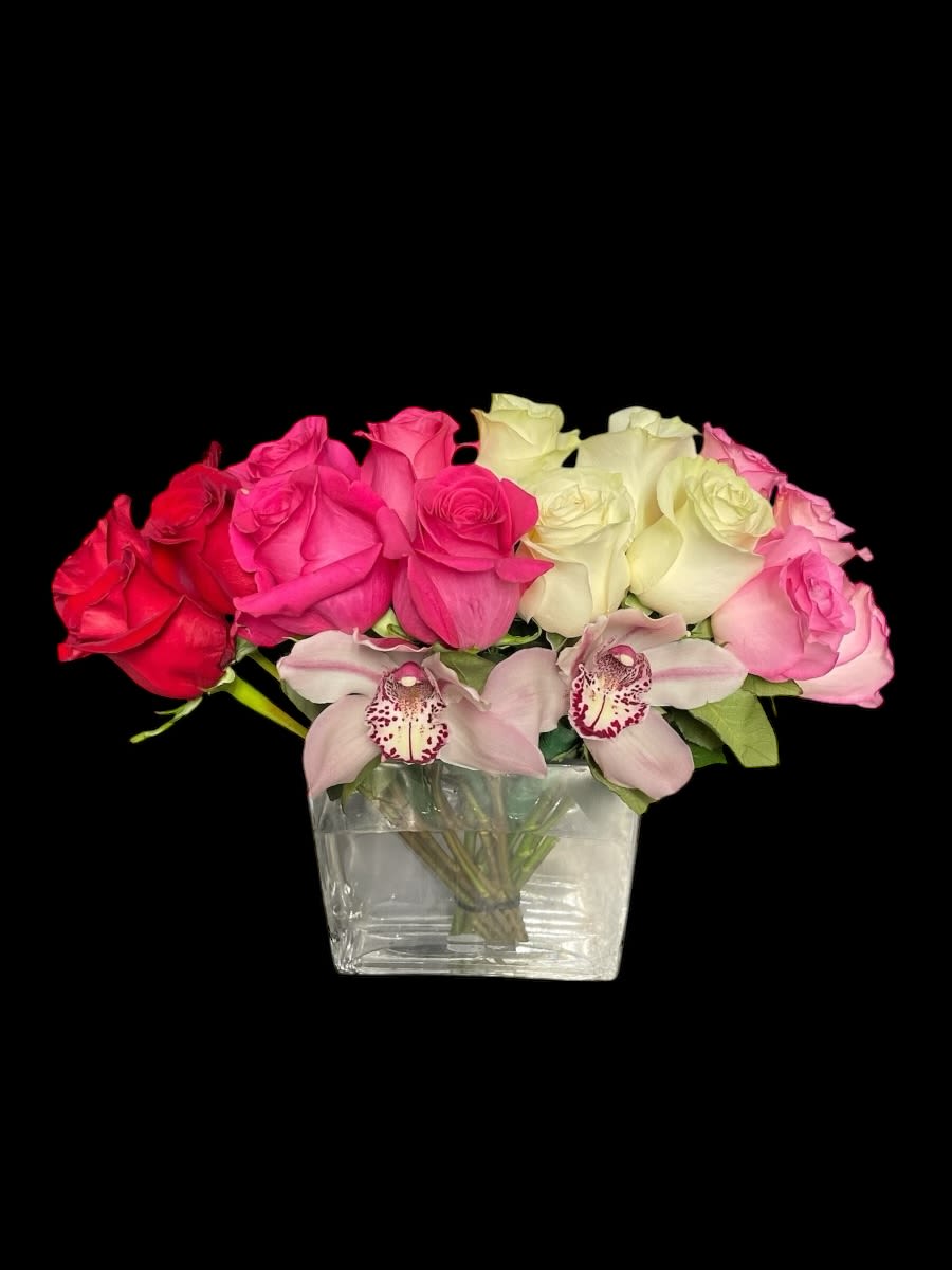 Low arrangement of red, pink, and ivory roses with two pink cymbidium orchids in a clear glass cube vase.