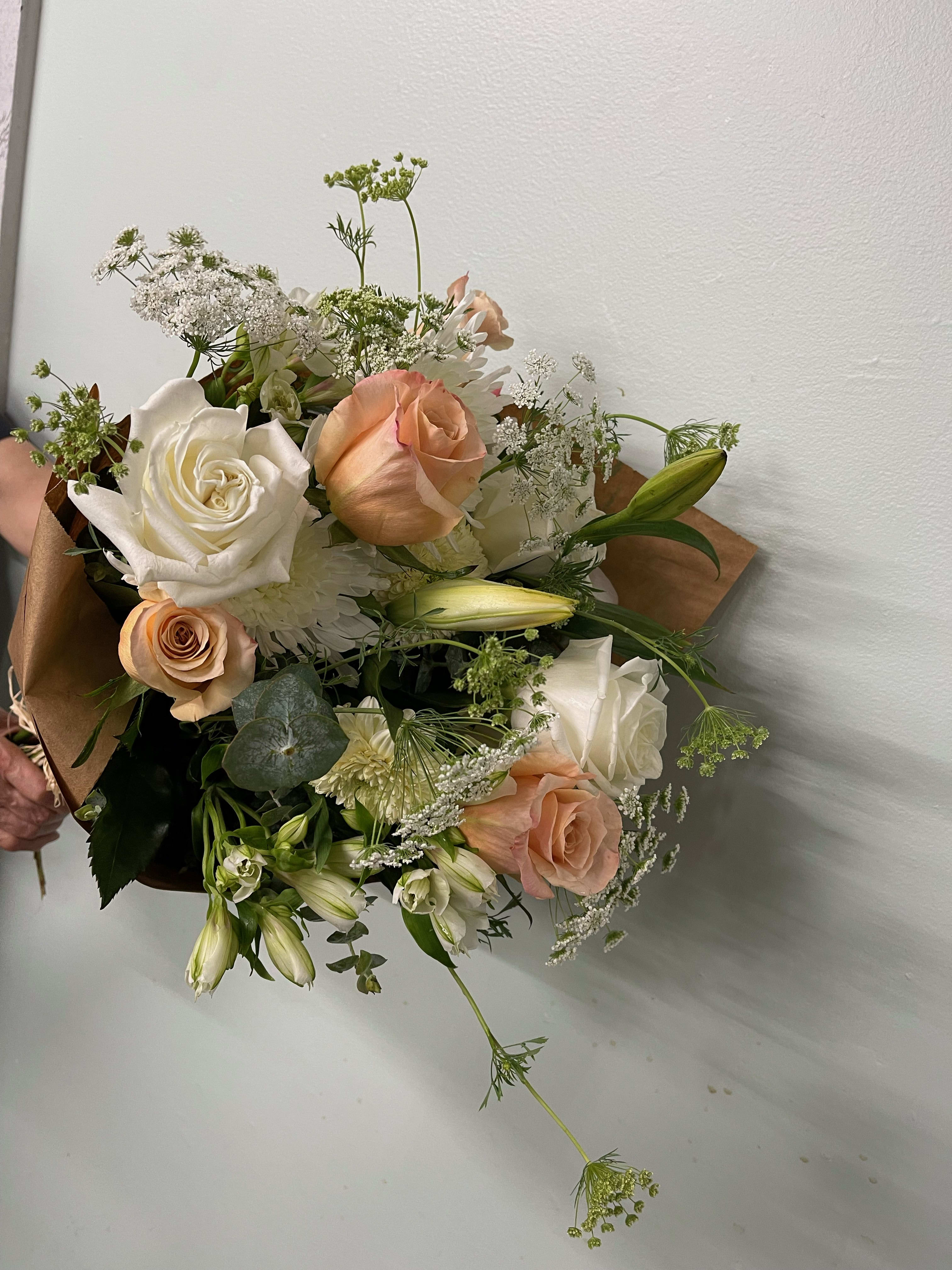 Hand-tied bouquet of white and peach roses with lilies and delicate white filler flowers in kraft paper wrap.