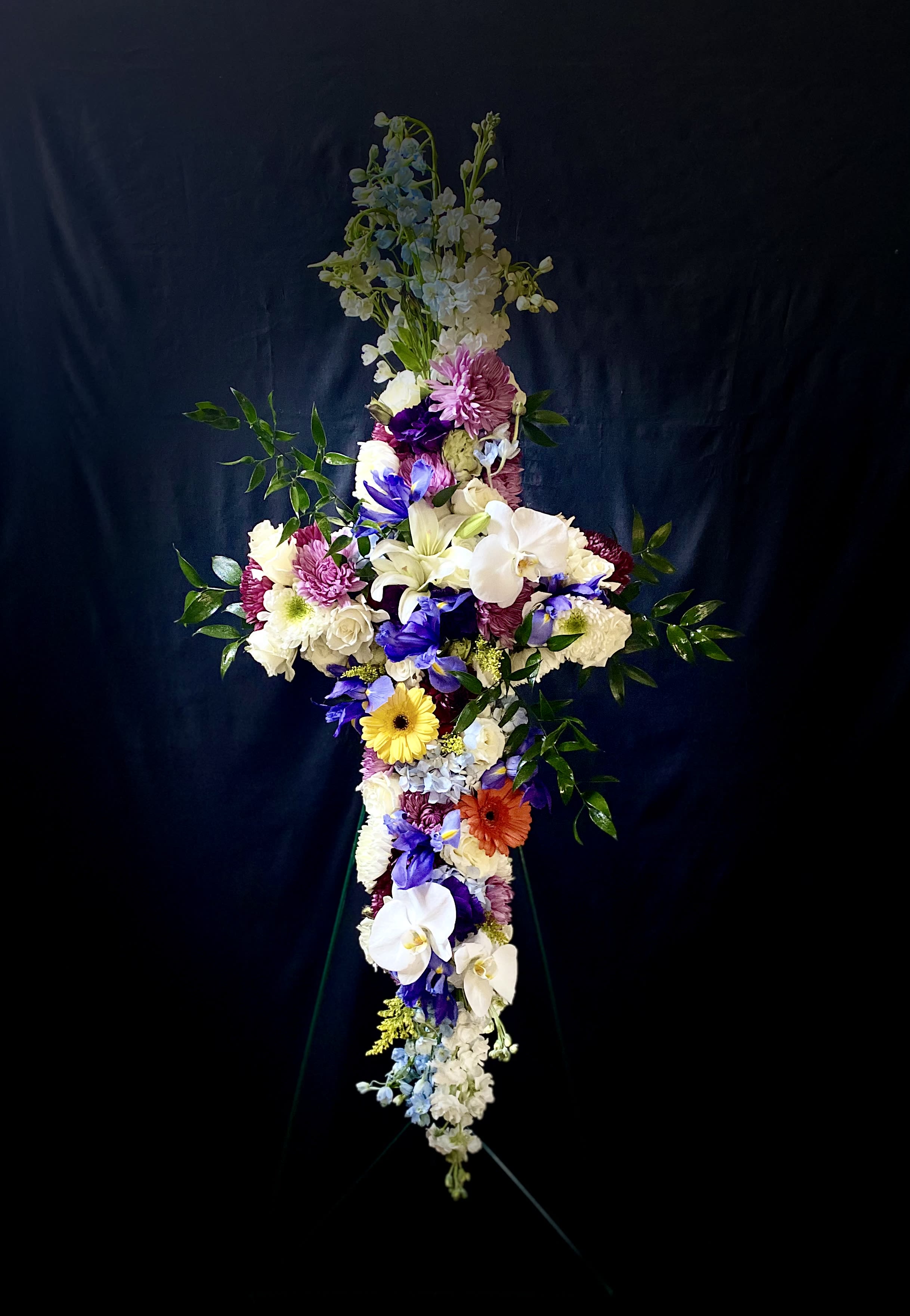 Tall cross-shaped standing spray with white lilies, orchids, mums and colorful daisies on a metal easel