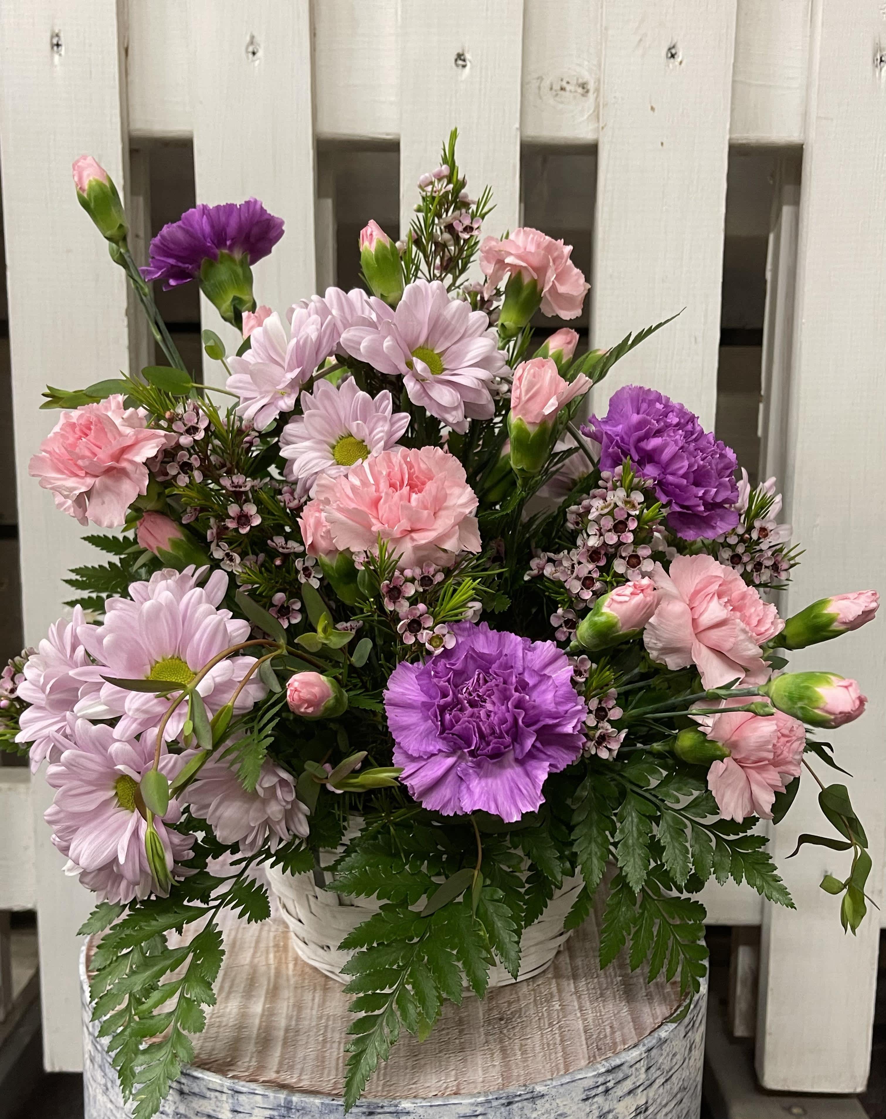 Basket arrangement of pink carnations, lavender daisies, and purple blooms in a white woven basket