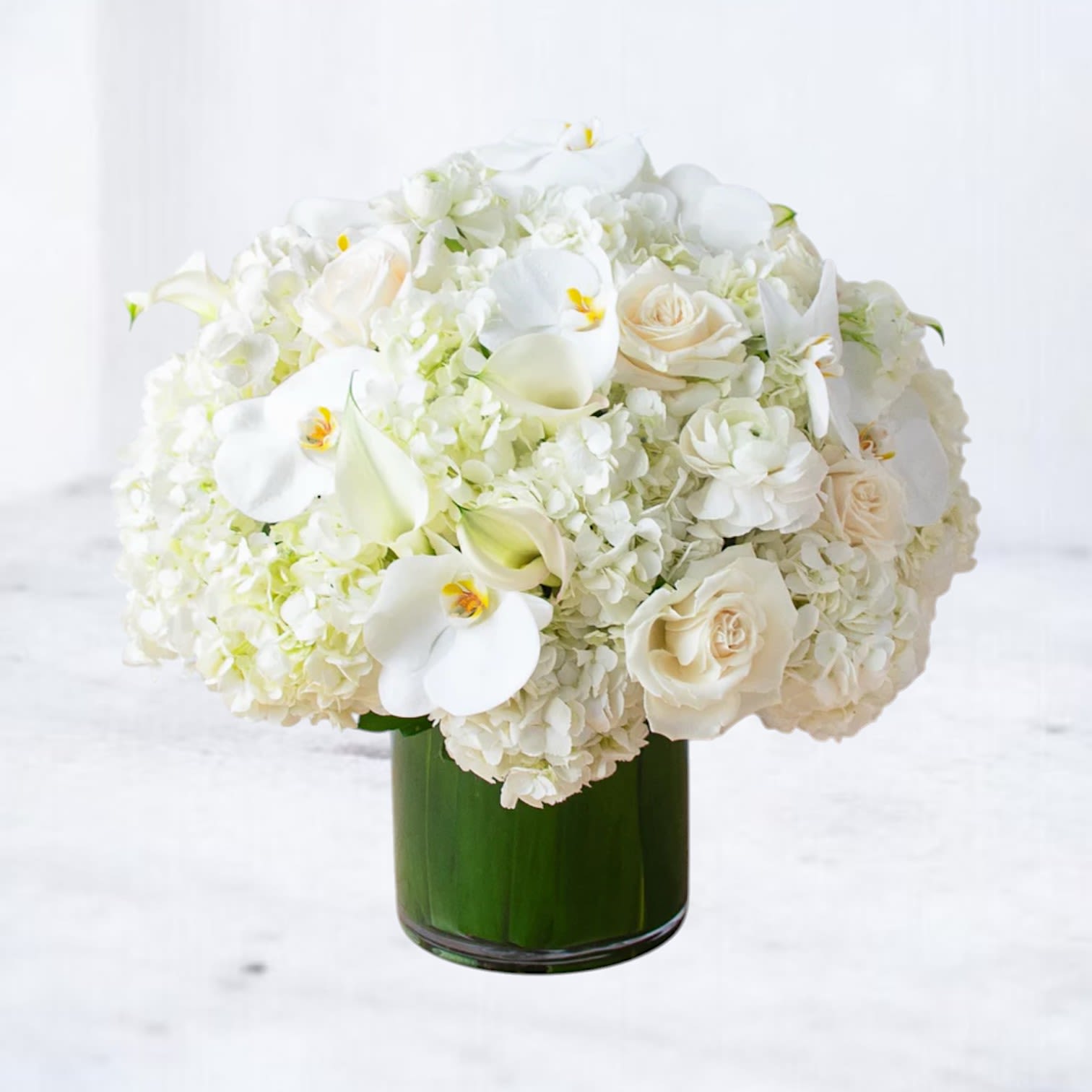 Time Stands Still Bouquet - The Time Stands Still Bouquet is a breathtaking expression of elegance and grace, perfect for commemorating cherished memories, celebrating everlasting love, or offering heartfelt condolences. Featuring a lush arrangement of white hydrangeas, delicate calla lilies, soft ivory roses, and exquisite white orchids, this bouquet radiates purity and sophistication.  Artfully arranged in a modern glass vase with a leaf-lined base, this design creates a serene atmosphere that brings comfort and beauty to any occasion. Whether marking a milestone anniversary, or celebrating a love that transcends time, the Time Stands Still Bouquet delivers a message of peace and enduring devotion.