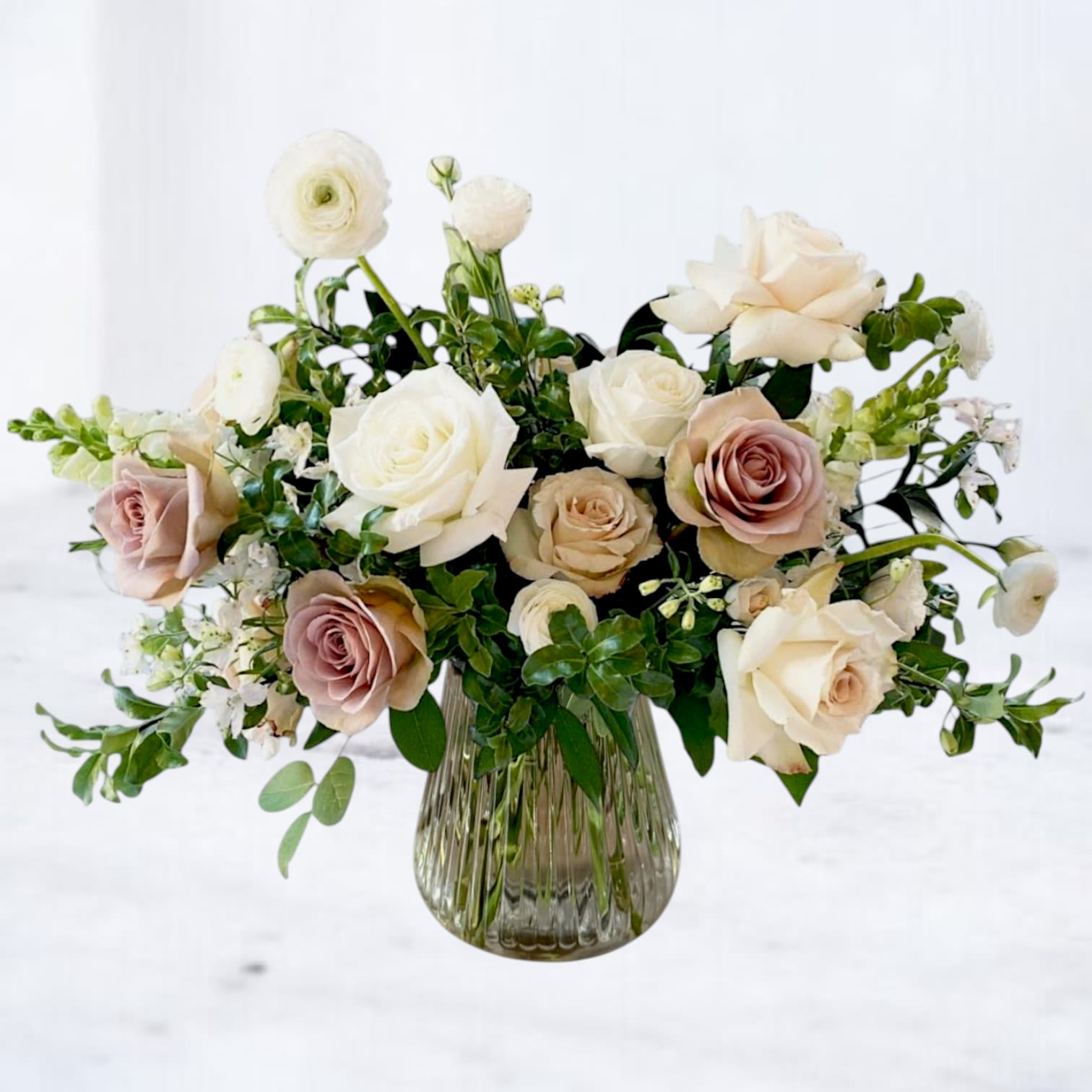 Mink Glow - A soft cascade of white and blush blooms creates an intimate, romantic feeling that's perfect for an anniversary bouquet or an "I'm sorry" gesture. This mixed-color flower arrangement combines lisianthus, ranunculus, roses, and snapdragons in white, peach, and pink tones so the person you're thinking of feels truly seen-gentle, sincere, and comforted.  Each bloom is spaced to showcase its shape, with creamy white ranunculus and blush roses taking center stage and delicate lisianthus and snapdragon accents adding light texture and movement. Smaller white buds weave through the composition for airiness, and the stems sit in a tall ribbed clear glass vase that gives the design a refined, understated finish while letting the soft palette glow.  Ideal for love and romance or to make amends, this arrangement suits both celebratory and restorative moments-the kind of bouquet that quiets the room and lifts the heart. Available in a tall presentation; easy care includes fresh water in the vase and a fresh cut before display to extend bloom life.