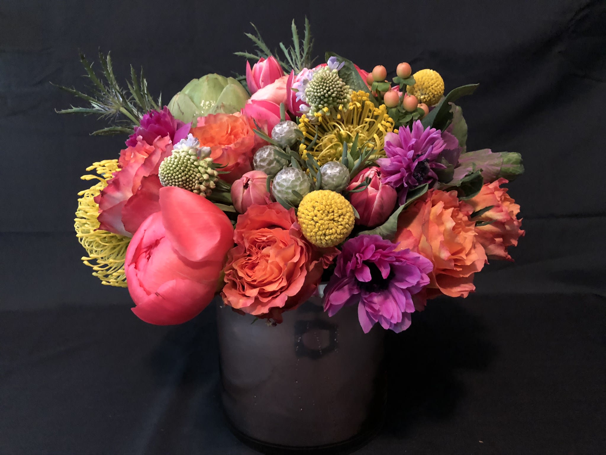 Bright mixed arrangement of pink, orange, yellow, and purple flowers in a smoky glass cylinder vase