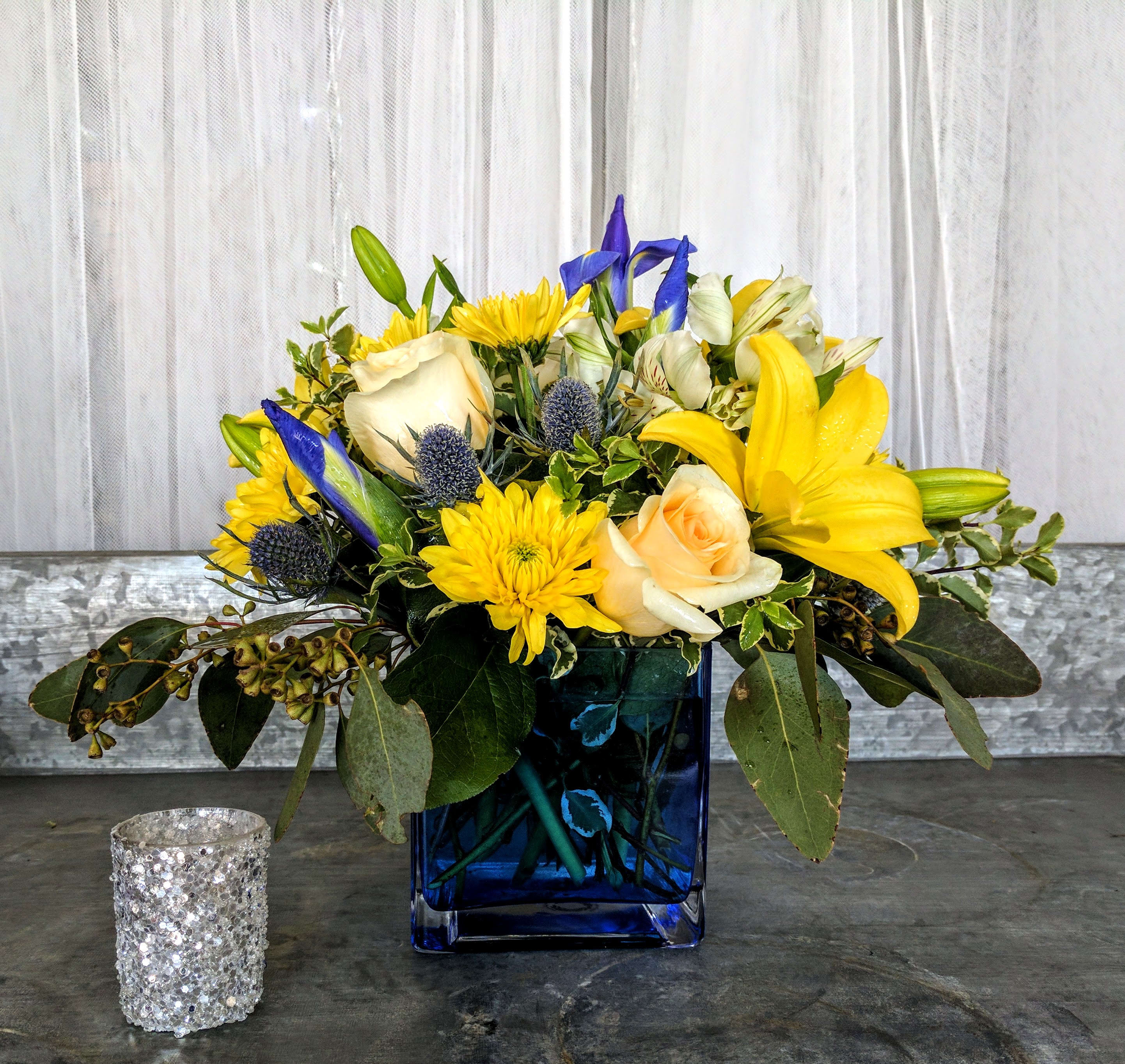 Compact arrangement of yellow lilies, cream roses, and blue accents in a blue glass cube vase.