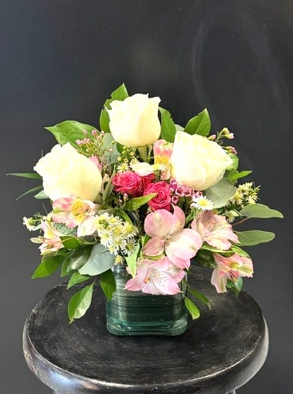#A127 - Sugar Cube - Soft pink spray roses and alstroemeria cradle three tall white roses to create a calm, uplifting centerpiece ideal for a birthday bouquet, Mother's Day gift, or a fresh get-well surprise. This fresh flower arrangement for occasions like anniversaries, new baby visits, or a simple "thinking of you" says gentle affection and quiet celebration-the kind of gift that makes the person you're thinking of feel truly seen. Blended with small white daisy-like chrysanthemums and lush green foliage, the blossoms are tightly clustered for a full, rounded silhouette that reads both elegant and casual.   The compact design sits in a low green glass cube vase, perfect for tabletops, desks, or a kitchen counter; the vase's clean lines let the pink, white, and green palette take center stage. Light, garden-fresh textures and a balanced scale make this a versatile choice for Easter, springtime moments, graduations, housewarmings, or to say "I'm sorry" or "thank you" with warmth.