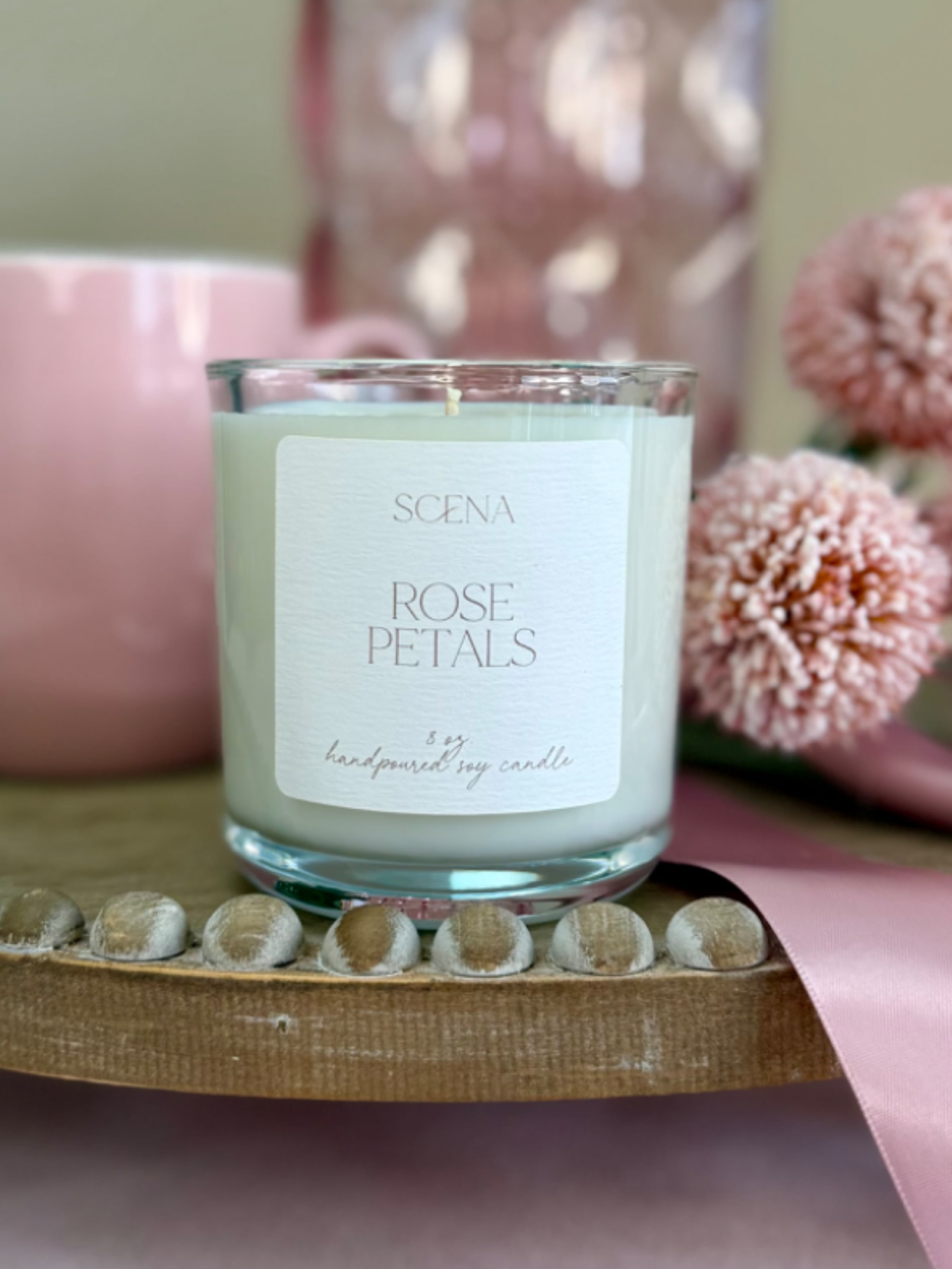 Rose-scented white candle in a clear glass jar with pink mug, flowers, and ribbon on a wooden tray
