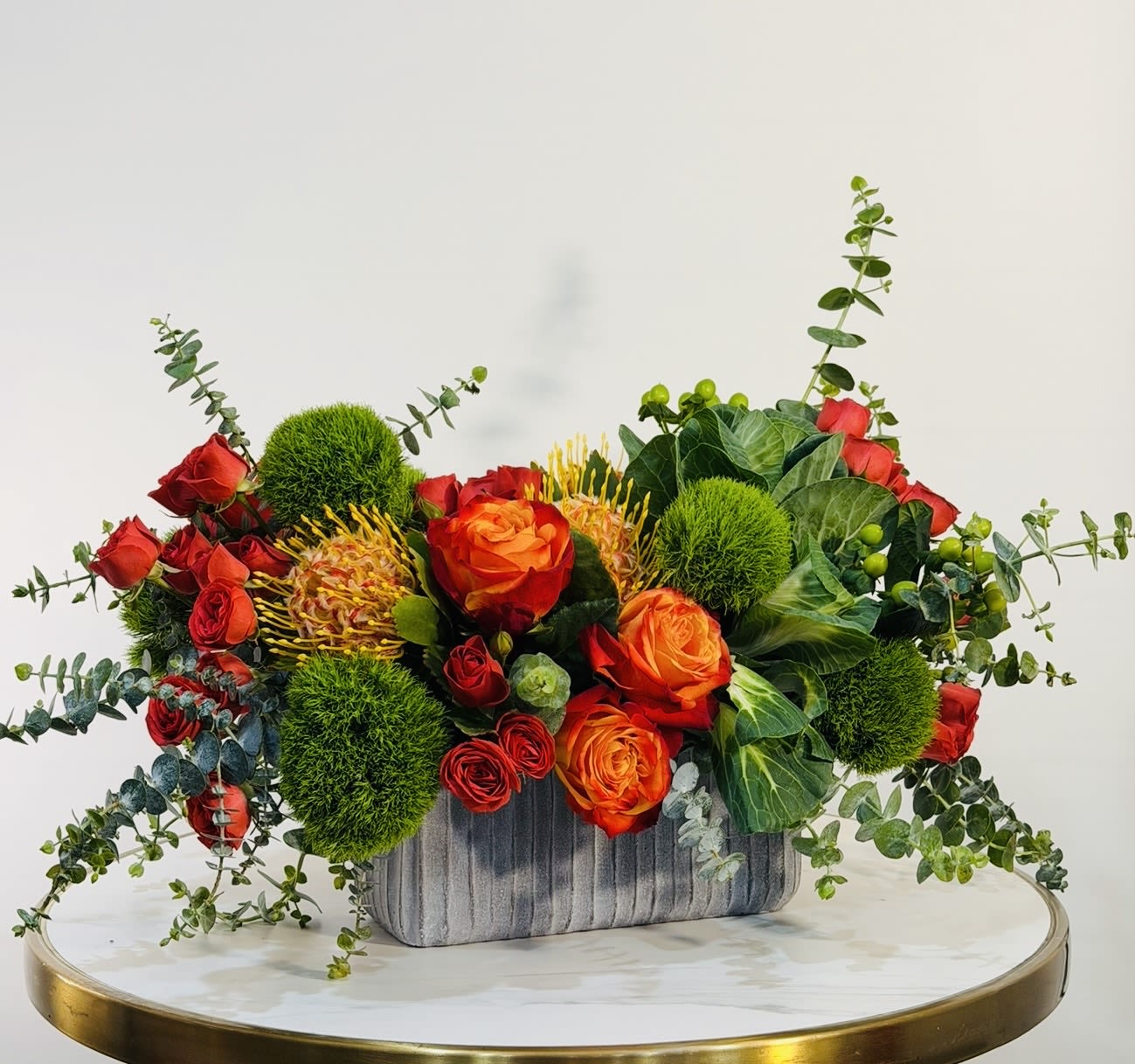 Sunset Fire - A stunning and vibrant floral arrangement featuring rich red and fiery orange roses, textured green elements, and unique seasonal accents arranged in a modern ceramic vase. This design brings warmth, energy, and elegance into any space, making it perfect for celebrations, gifts, or simply making a statement. Each arrangement is handcrafted with care using fresh, premium flowers to ensure long lasting beauty and impact. Same Day Flower Delivery Available across Beverly Hills, Bel-Air, West Hollywood, Beverly Grove, Santa Monica, Culver City, Hollywood Hills, Hollywood, Studio City, Encino, and Sherman Oaks.