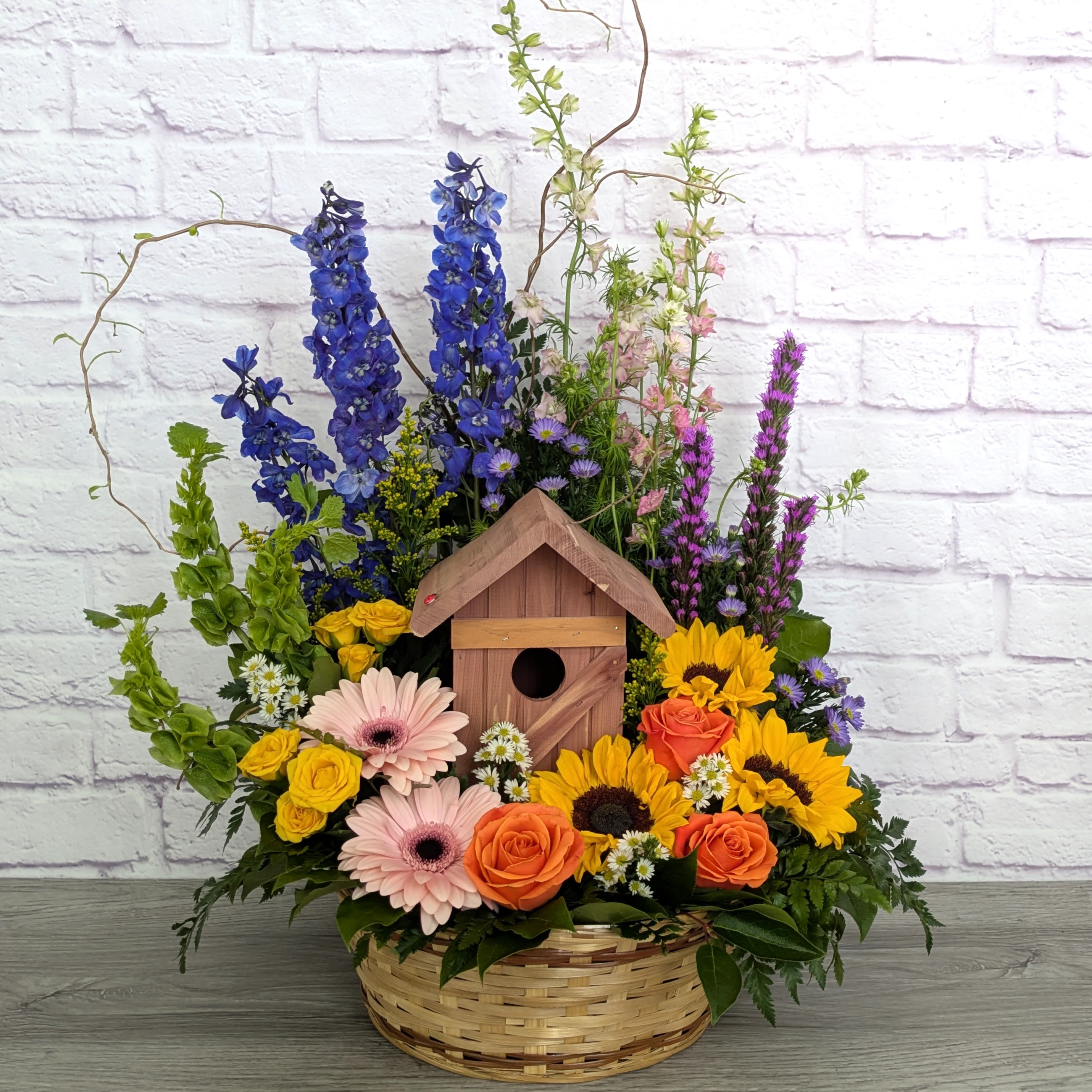 Tina's Bird House - Tina's Bird House is A Ladybug Floral exclusive bouquet. This bouquet includes curly willow, larkspur, delphinium, bells of ireland, liatris, sunflower, gerberadaisies, roses, spray rose, solidago and monte Casino. Featured in a 12” wicker basket with a functional wooden bird house.  Orientation: All Around 28” Tall 22” Wide   In honor of Tina 1966 - 2026 