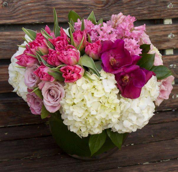 Low round bouquet of white hydrangeas, pink roses and tulips, and purple orchids in a clear glass vase