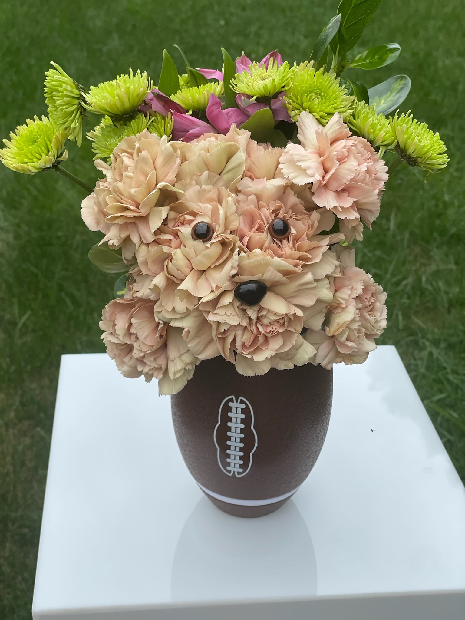 Dog-shaped peach carnation arrangement with green mums in a brown football vase on a white stand.