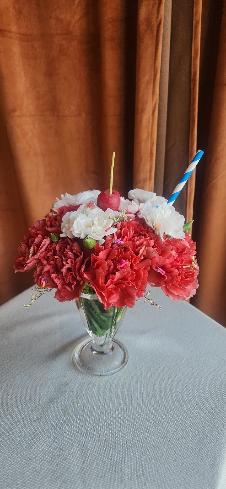 Cherry on Top  - A sweet twist on florals, Cherry on Top is our playful take on a classic ice cream sundae reimagined through blooms. Designed in a charming glass dish, this arrangement features ruffled, scoop-like carnations in rich cherry reds and soft vanilla whites, finished with whimsical details that bring it all to life. 