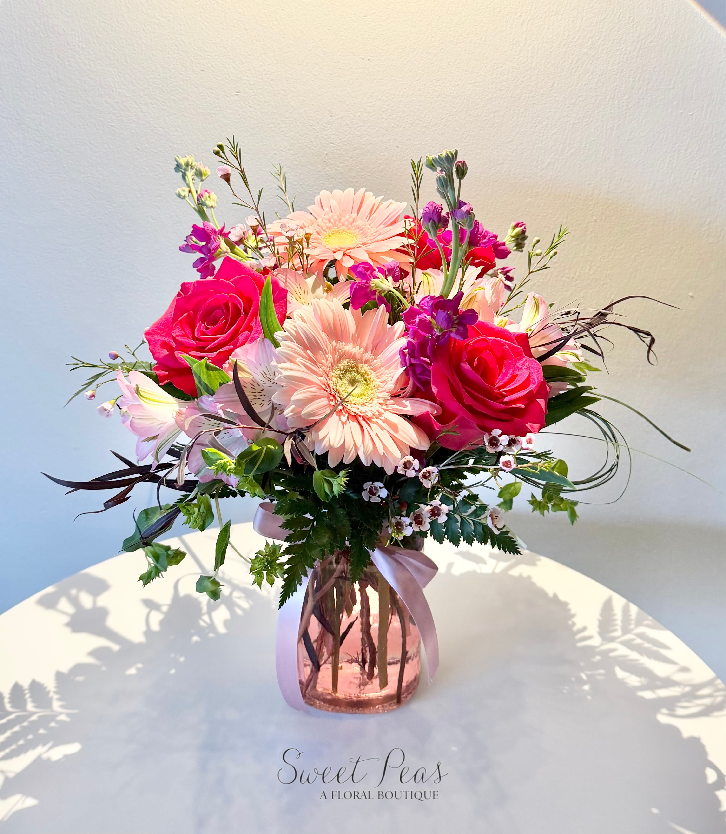 Pink roses and pale gerbera daisies arranged with mixed blooms in a pink glass vase with ribbon.