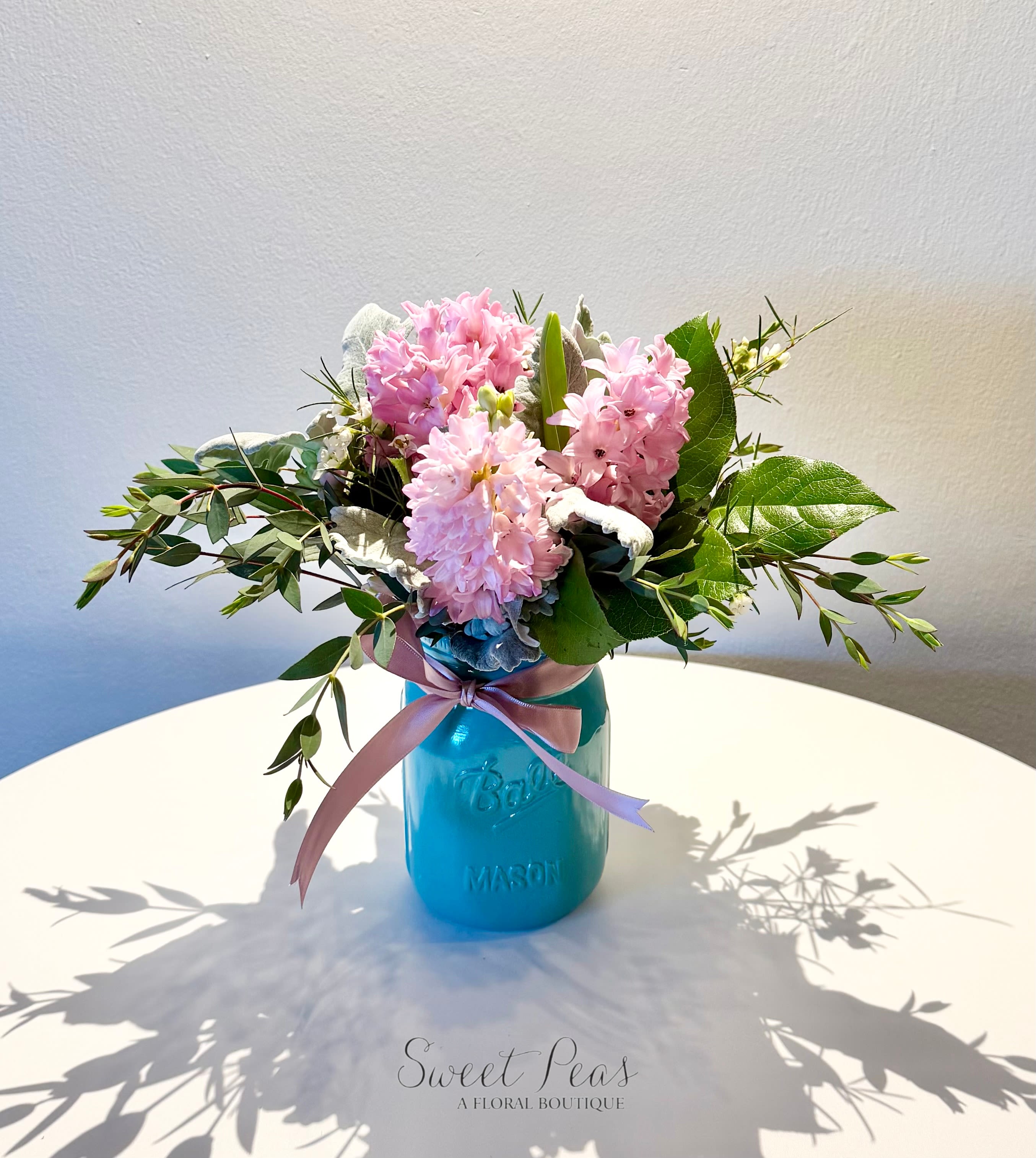 Hyacinth Breeze - Delicately gathered stems of fragrant hyacinth create a lush, textural design that celebrates the beauty of a single bloom. Hyacinth Breeze is arranged with natural greenery in a vibrant mason jar and finished with ribbon for a fresh, garden-inspired look. Known for its sweet, unmistakable scent, this arrangement is perfect for brightening a space and filling it with the essence of spring.  As Shown has five hyacinth stems; Deluxe Version has 7 Hyacinth Stems; Premium has 10 Hyacinth Stems