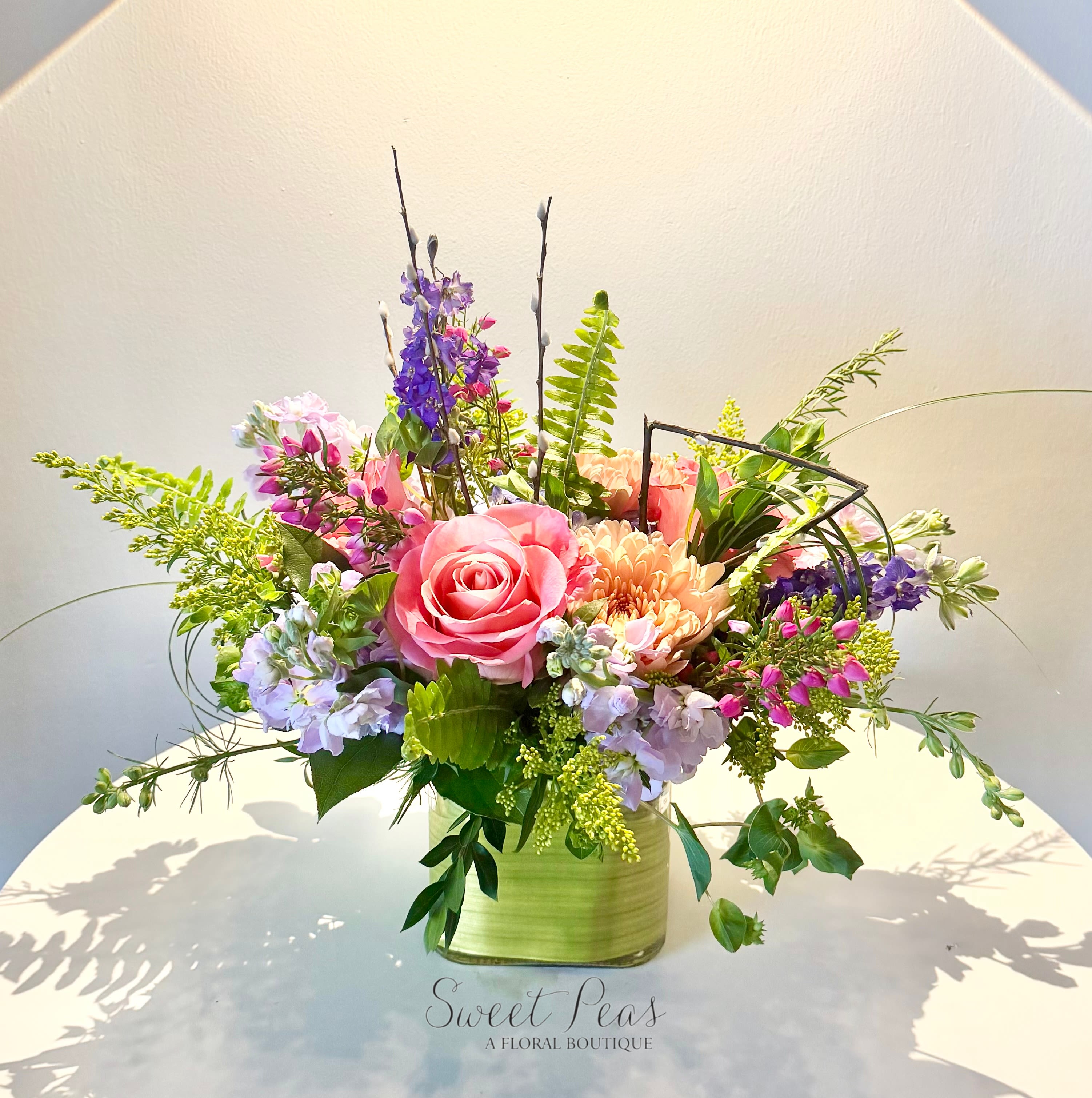 Garden Muse - Playful and artistic, Garden Muse is a vibrant, garden-style arrangement bursting with fresh color and texture. Featuring a lively mix of pinks, peaches, purples, and soft greens, this design is accented with natural greenery and a modern geometric detail for a unique, contemporary touch. Designed in a leaf wrapped vessel, it’s a cheerful and creative piece perfect for brightening someone’s day or celebrating any occasion.