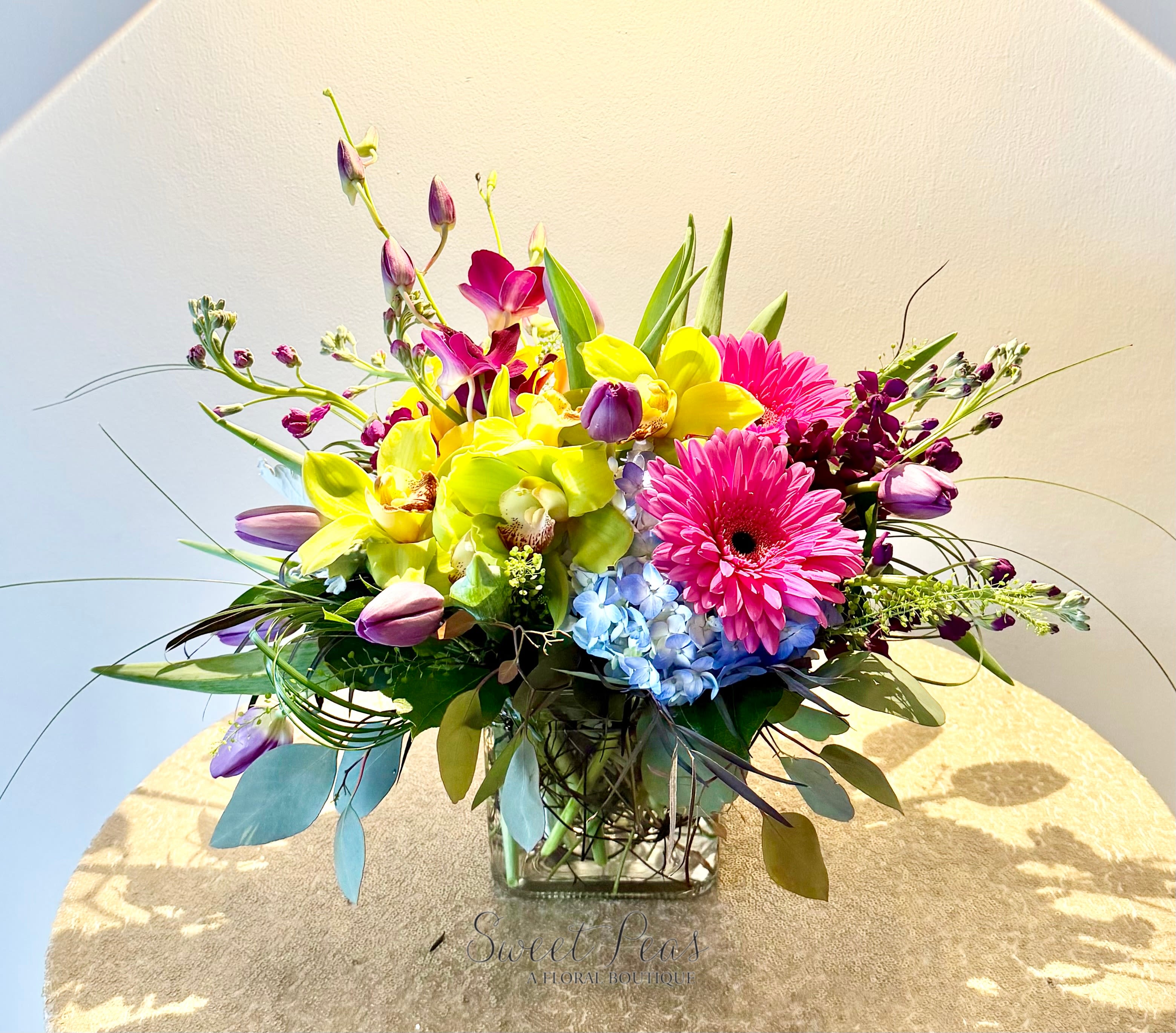 Low mixed arrangement of pink gerbera daisies, yellow orchids, tulips, and blue hydrangea in a clear glass cube vase