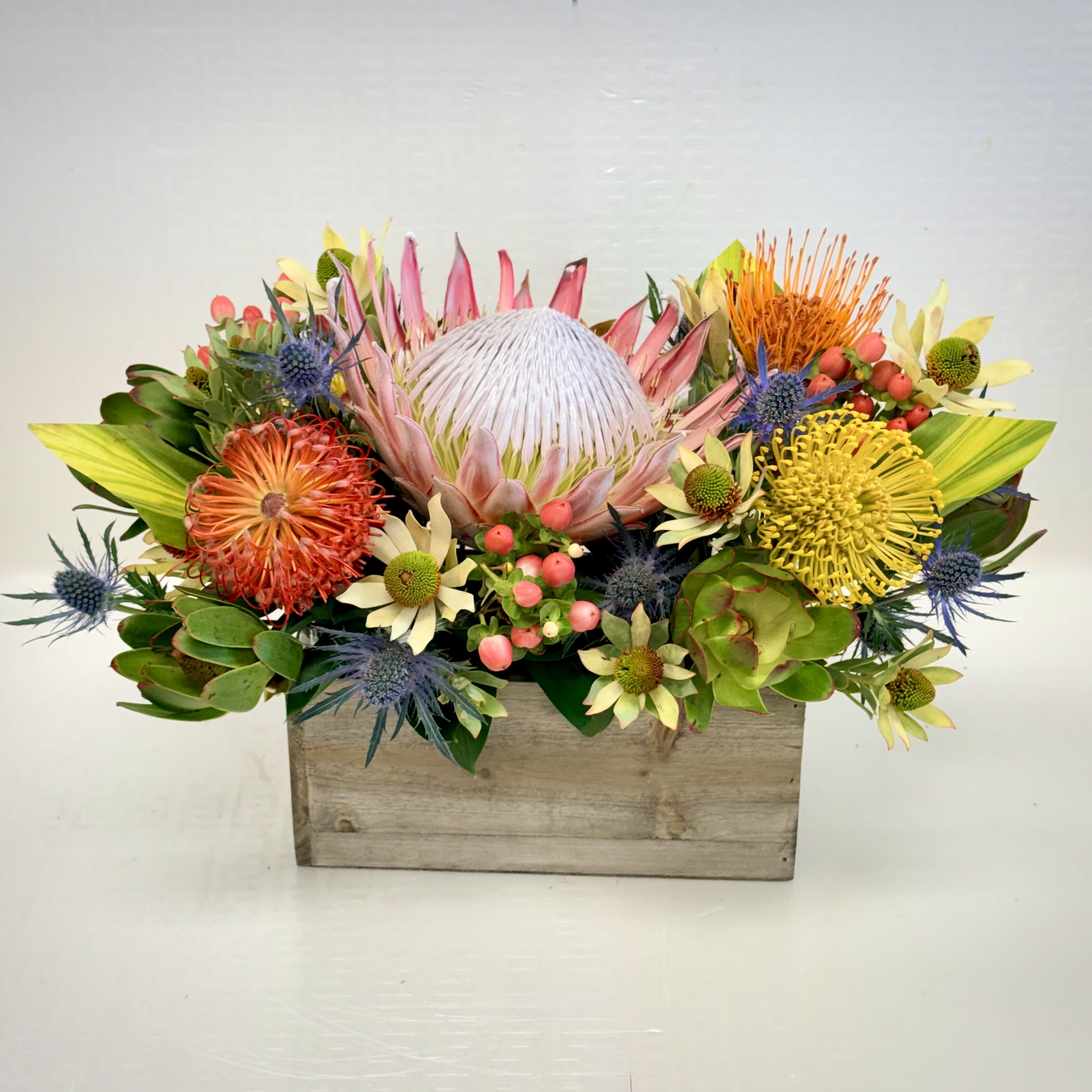 Protea Paradise Garden - A bold and cheerful arrangement centered around a magnificent king protea, complemented by vibrant seasonal blooms and textured greenery in a rustic wooden container. Perfect for brightening celebrations, sending warm wishes, or making a memorable statement gift.