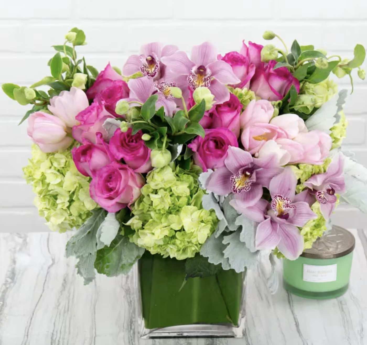 Royal Garden (DB) - Indulge in the charm of our modern floral arrangement, featuring a captivating blend of green hydrangeas, pink roses, pink tulips, and pink cymbidium orchids. Expertly arranged in a large leaf-lined glass cylinder vase, this ensemble combines vibrant colors, varied textures, and natural beauty.  At its heart, lush green hydrangeas provide a lively backdrop for the soft, velvety pink roses that add elegance and romance with their sweet fragrance. Delicate pink tulips bring freshness and playfulness, while striking pink cymbidium orchids contribute exotic sophistication.  The leaf-lined glass vase offers a contemporary base, its transparency highlighting each bloom’s beauty. Its large size makes this arrangement a bold statement piece, ideal for enhancing living rooms, entryways, or event venues.  Elevate your space with this stunning floral display that captivates the senses. Perfect as a statement piece or a gift, it radiates elegance, romance, and modern sophistication.