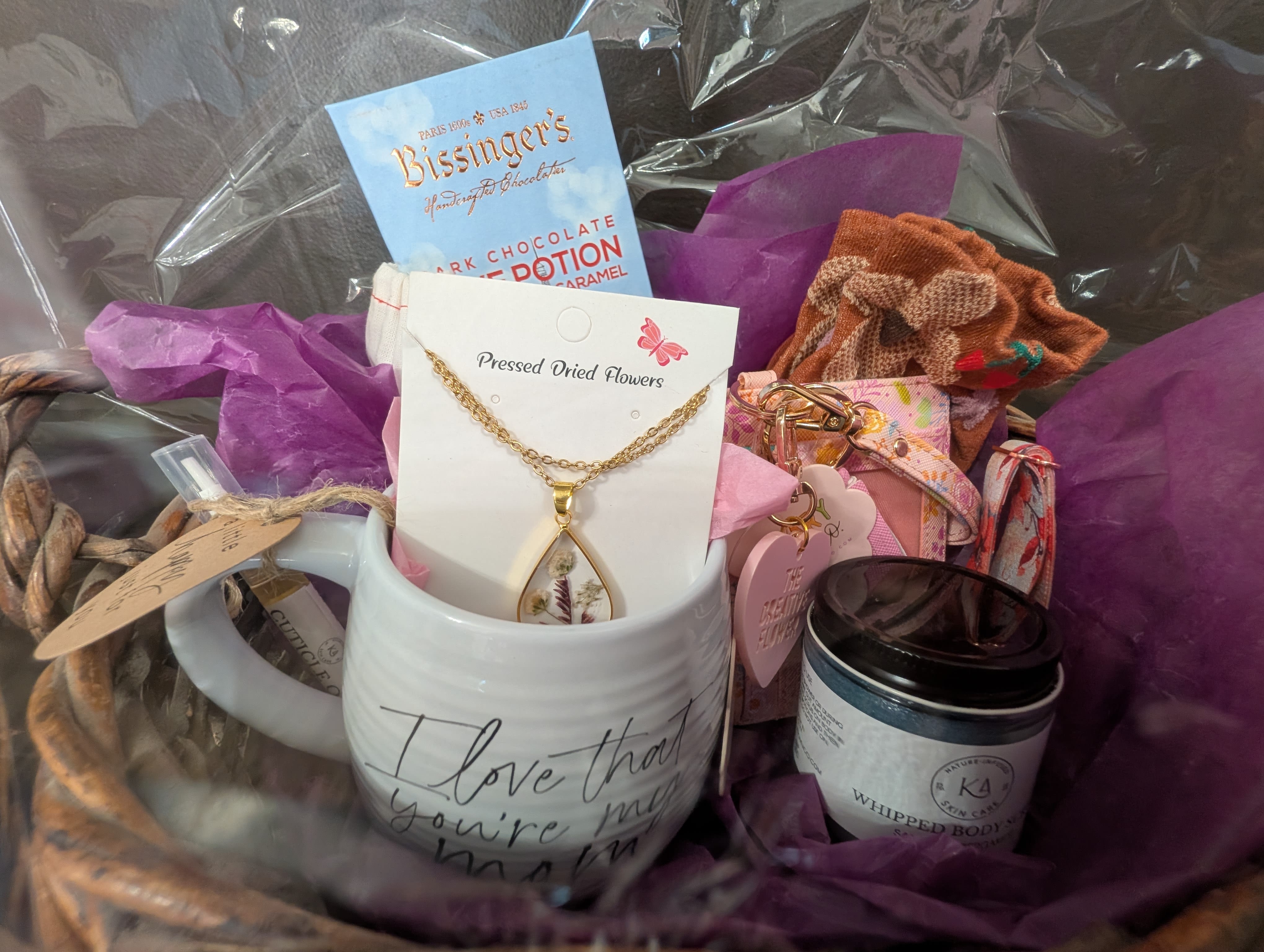 Mother's Day Gift Basket - A gift basket for moms, with a mug, socks, chocolate, body scrub, pressed flower necklace, wallet, and cuticle oil.