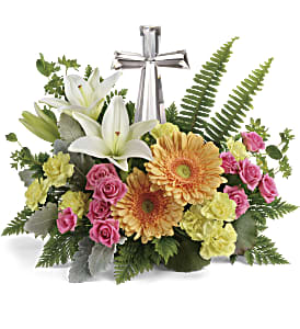 Low arrangement of white lilies, peach gerbera daisies, pink roses and yellow carnations with a silver cross.