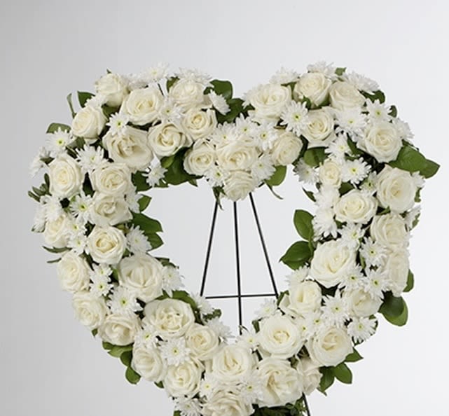 PURE DEVOTION FUNERAL HEART - A graceful expression of everlasting love, the Pure Devotion Heart is designed with lush white roses, delicate white seasonal flowers, and fresh greenery in a timeless open-heart silhouette. Each bloom is thoughtfully placed to symbolize peace, remembrance, and the unbreakable bond shared with a loved one. This elegant sympathy tribute offers a serene and heartfelt way to honor a life beautifully lived. The soft whites convey purity and reverence, while the natural textures bring a sense of comfort during difficult moments. Handcrafted with premium blooms and intentional design, this standing heart is a meaningful centerpiece for funeral or memorial services—offering a quiet, powerful message of love that lives on.