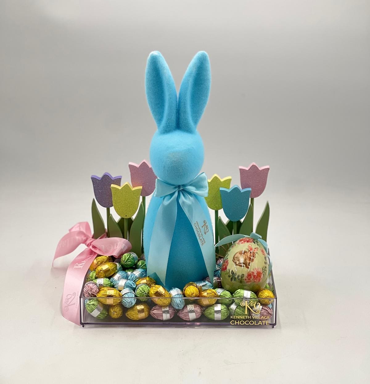Easter Bunny Chocolate Garden - A playful and vibrant Easter display, this charming arrangement features a soft blue bunny centerpiece surrounded by a bed of premium wrapped chocolates in cheerful pastel colors. Accented with colorful tulip-inspired décor and a decorative Easter egg, the design brings a joyful springtime atmosphere to life. Finished with delicate ribbons and presented in a clear, modern tray, this piece is perfect for gifting or adding a festive touch to any Easter celebration.
