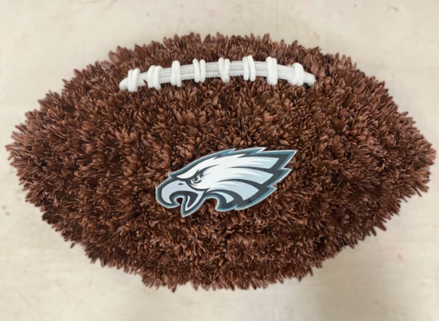 Eagles Football Tribute - Honor a loved one with a meaningful tribute inspired by their love for the Philadelphia Eagles. This listing features two custom floral designs: a football and a helmet, each carefully crafted to create a memorable and personalized display for funeral services or memorials. Both designs are made using fresh flowers and thoughtful detail, offering a unique way to celebrate a life and passion. Designs are approximately 18"-20".  Please note that the price listed is for one tribute only. You must choose either the football or the helmet design when placing your order.  These designs can also be created for other teams. To request a different team, please call or visit us in person to place your order.  If you would like a script included on a ribbon, please note your request in the florist note section when placing your order.  For delivery, please enter the name of the deceased in the recipient field and include the correct date of the service to ensure proper timing and placement.