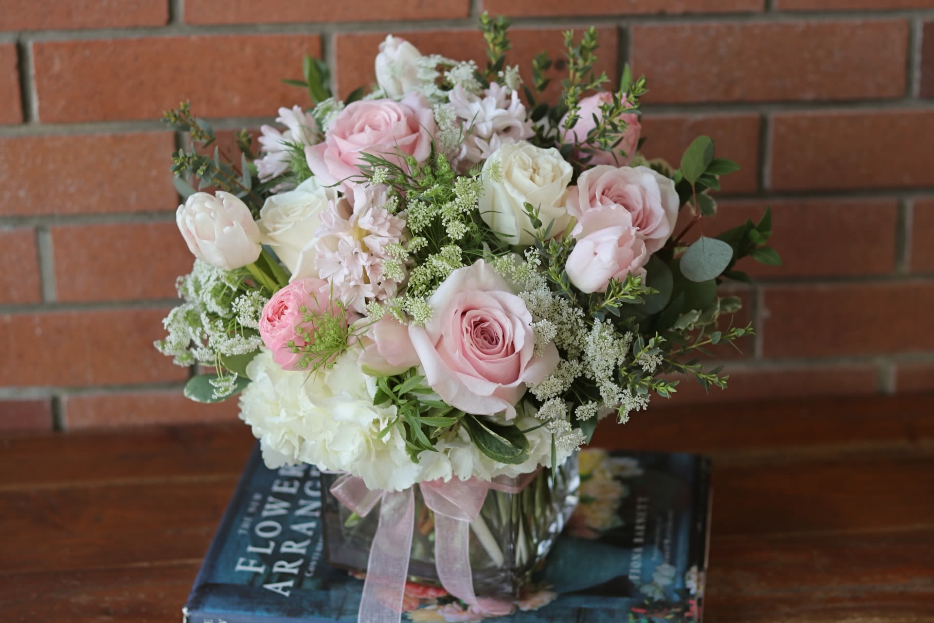 Low pink and white rose arrangement in a clear glass cube vase with ribbon, set on books.