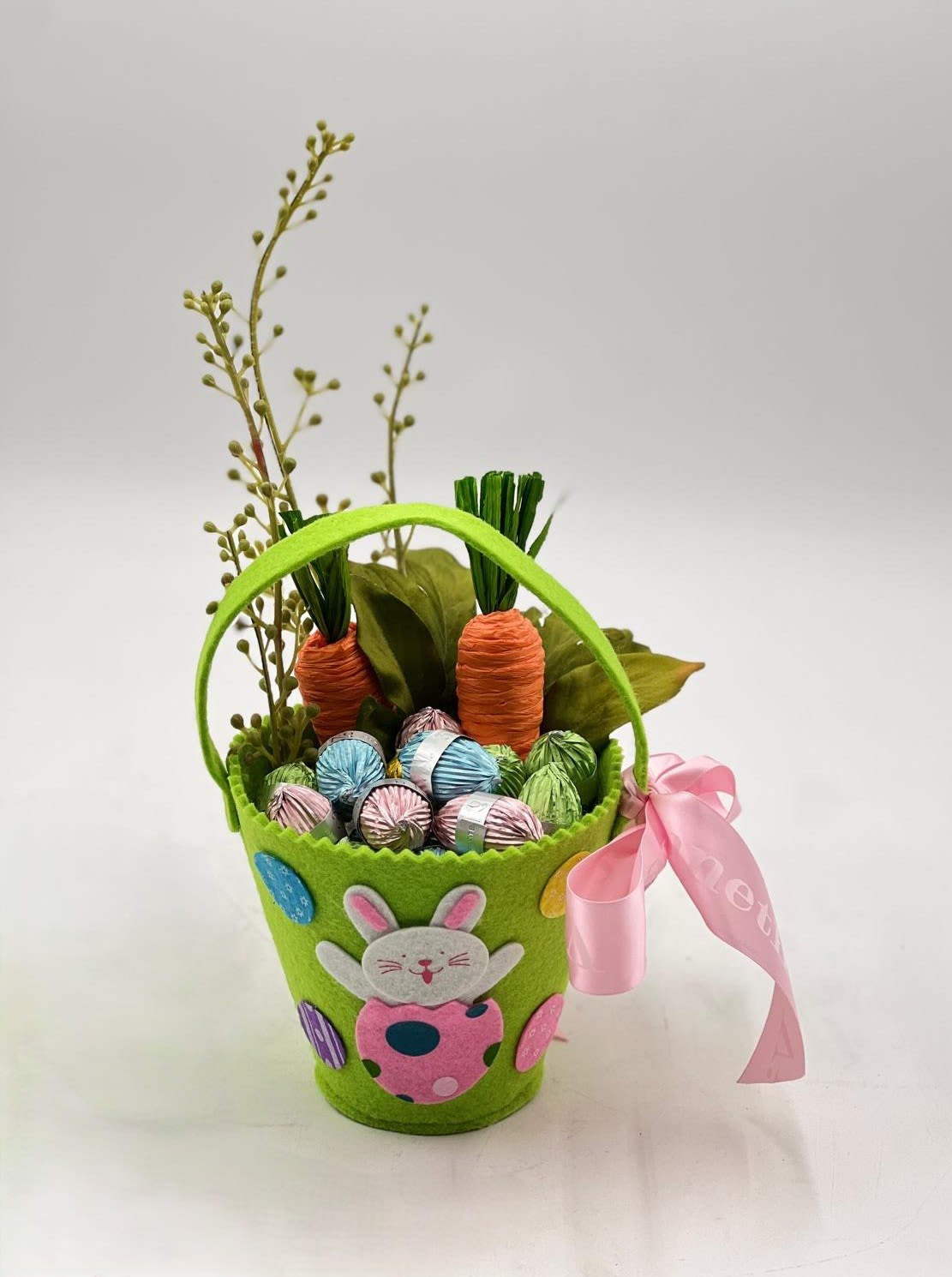 Carrot Garden Easter Chocolate Basket - A cheerful and playful Easter treat, this vibrant green basket is filled with premium wrapped chocolates in soft pastel colors, accented with whimsical carrot details and fresh greenery for a festive spring feel. The adorable bunny design on the basket adds a charming, childlike touch, while the soft pink ribbon brings a hint of sweetness and elegance. Perfect for gifting, this arrangement captures the joyful spirit of Easter in a fun and colorful presentation that delights both kids and adults.