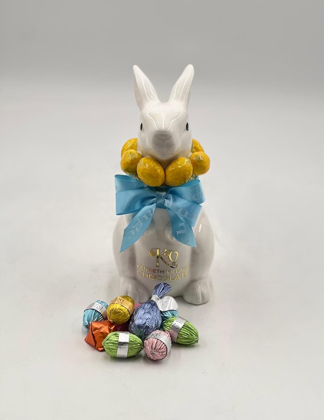 Elegant Easter Bunny Box - A refined and charming Easter keepsake, this elegant ceramic bunny container is adorned with a soft blue ribbon and a delicate ring of golden eggs around its neck, creating a timeless and festive look. Inside, it holds a selection of premium wrapped chocolates in cheerful pastel tones, making it both a decorative piece and a delightful treat. Perfect as a thoughtful gift or seasonal décor, this piece brings a touch of sophistication and sweetness to any Easter celebration.