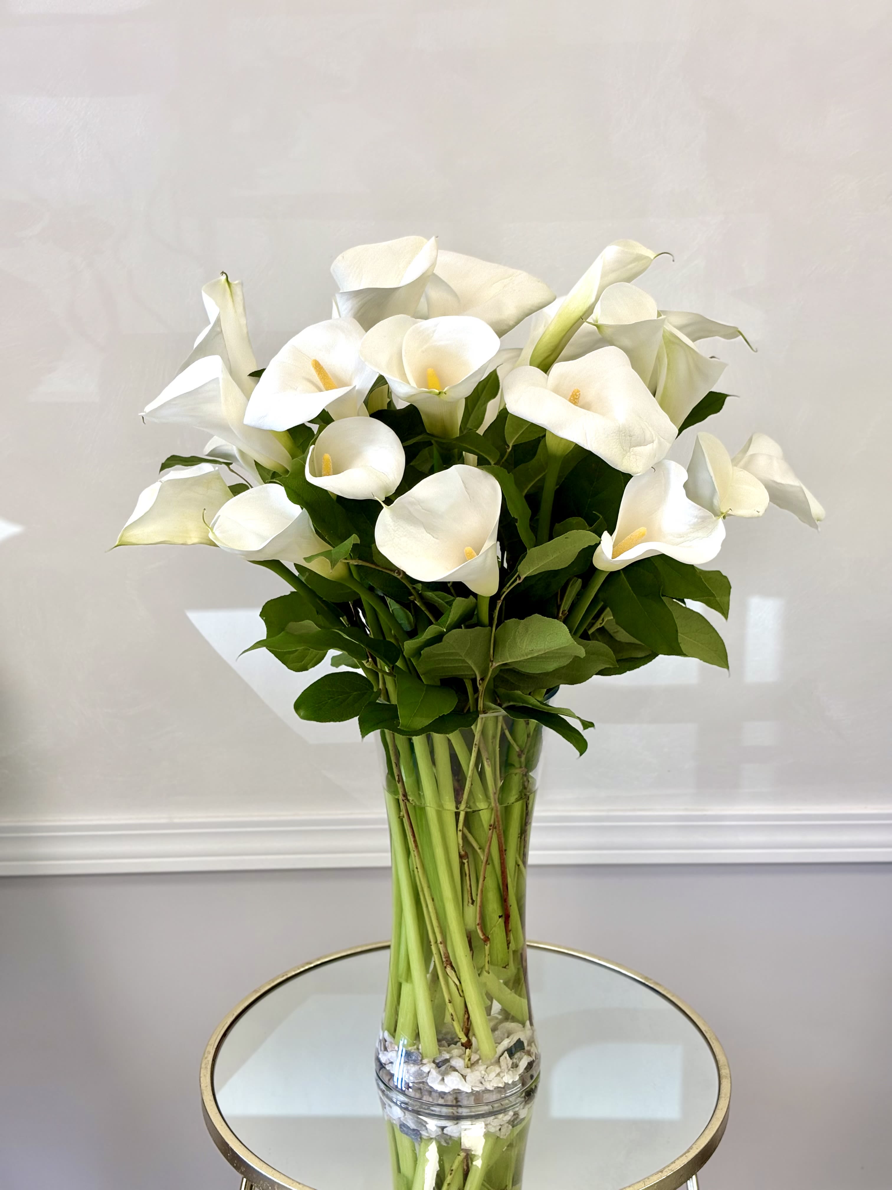 Calla Lily By VLM Flowers - Calla lilies are elegant, trumpet-shaped flowers known for their smooth, sculptural blooms and graceful appearance. Typically found in pure white with a yellow spadix center, they symbolize purity, sophistication, and beauty. Their sleek form and rich green foliage make them a popular choice for modern floral arrangements, weddings, and refined interior décor.