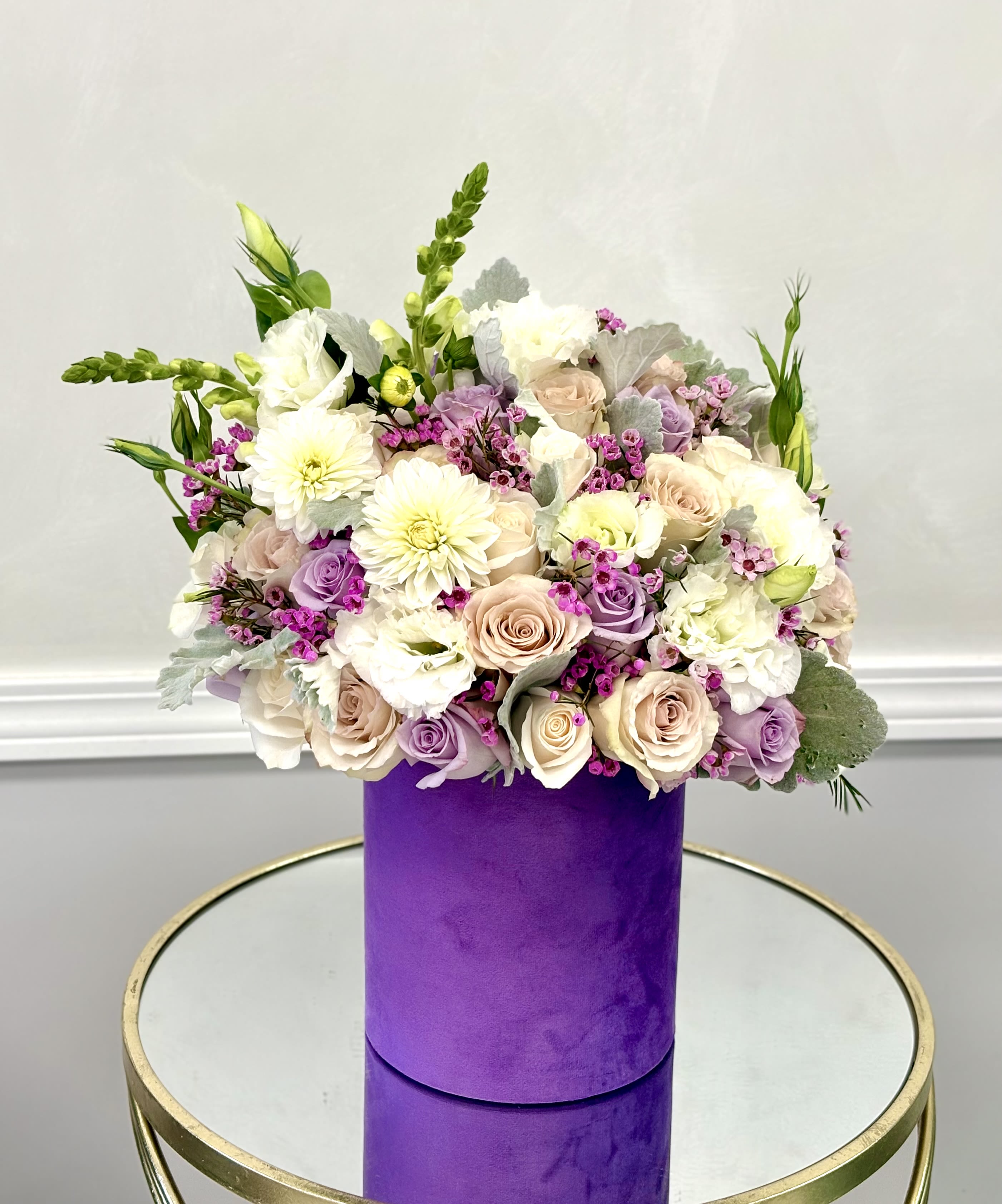 Lavender Whispers Hat Box By VLM Flowers - A graceful medley of soft lavender, blush, and cream blooms, Lavender Whispers is a floral daydream in a lilac hat box. Featuring lush roses, creamy dahlias, lisianthus, and waxflowers, this arrangement is a celebration of calm beauty and refined charm. Accents of dusty miller add a silvery softness, making it a perfect gift for anniversaries, sympathy gestures, or simply saying “you’re loved.”