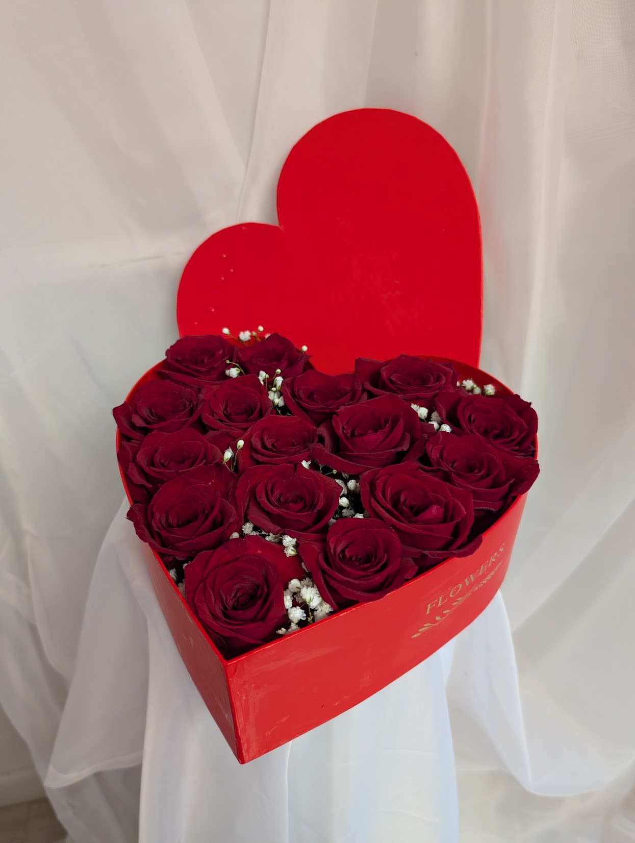 Heart-shaped red box filled with deep red roses and small white filler flowers