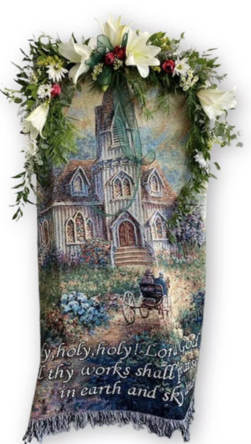 Religious tapestry blanket with church scene topped by white lilies and red roses with greenery.