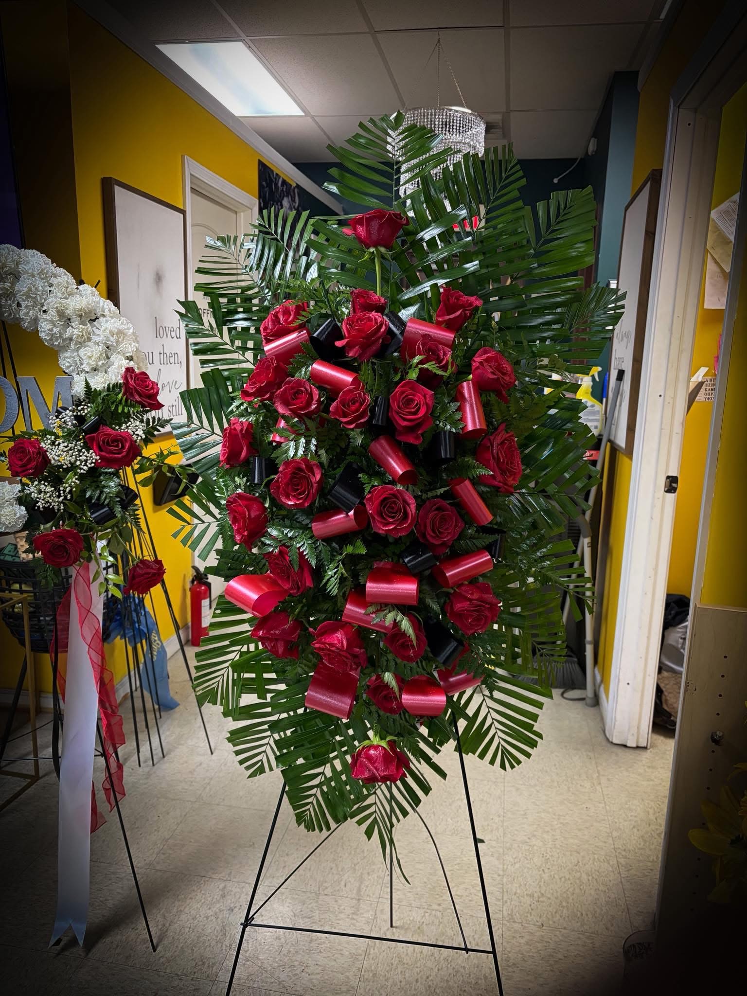 Red Remembrance - A large, elegant standing spray featuring rich red roses arranged in a full, oval shape. It’s accented with lush tropical greenery (like palm leaves) and decorative red and black ribbon loops, giving it a bold, dramatic presence. This type of arrangement is commonly used for funerals or memorial services and is displayed on an easel.  