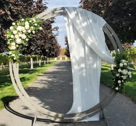 Tall circular metal arch with white flowers and draped white fabric on an outdoor path