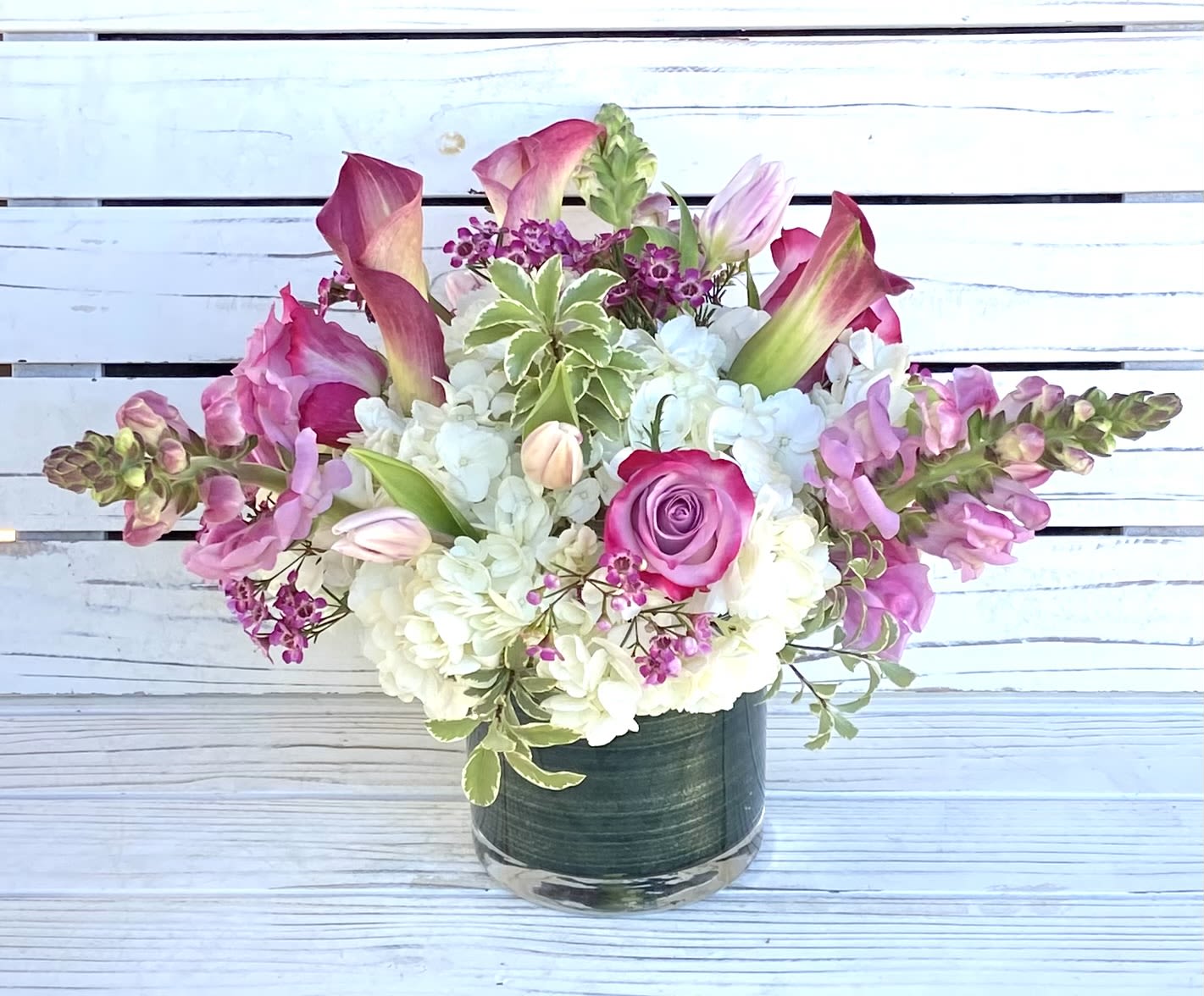 Pink and white flower arrangement with roses, calla lilies, and tulips in a glass cylinder vase.