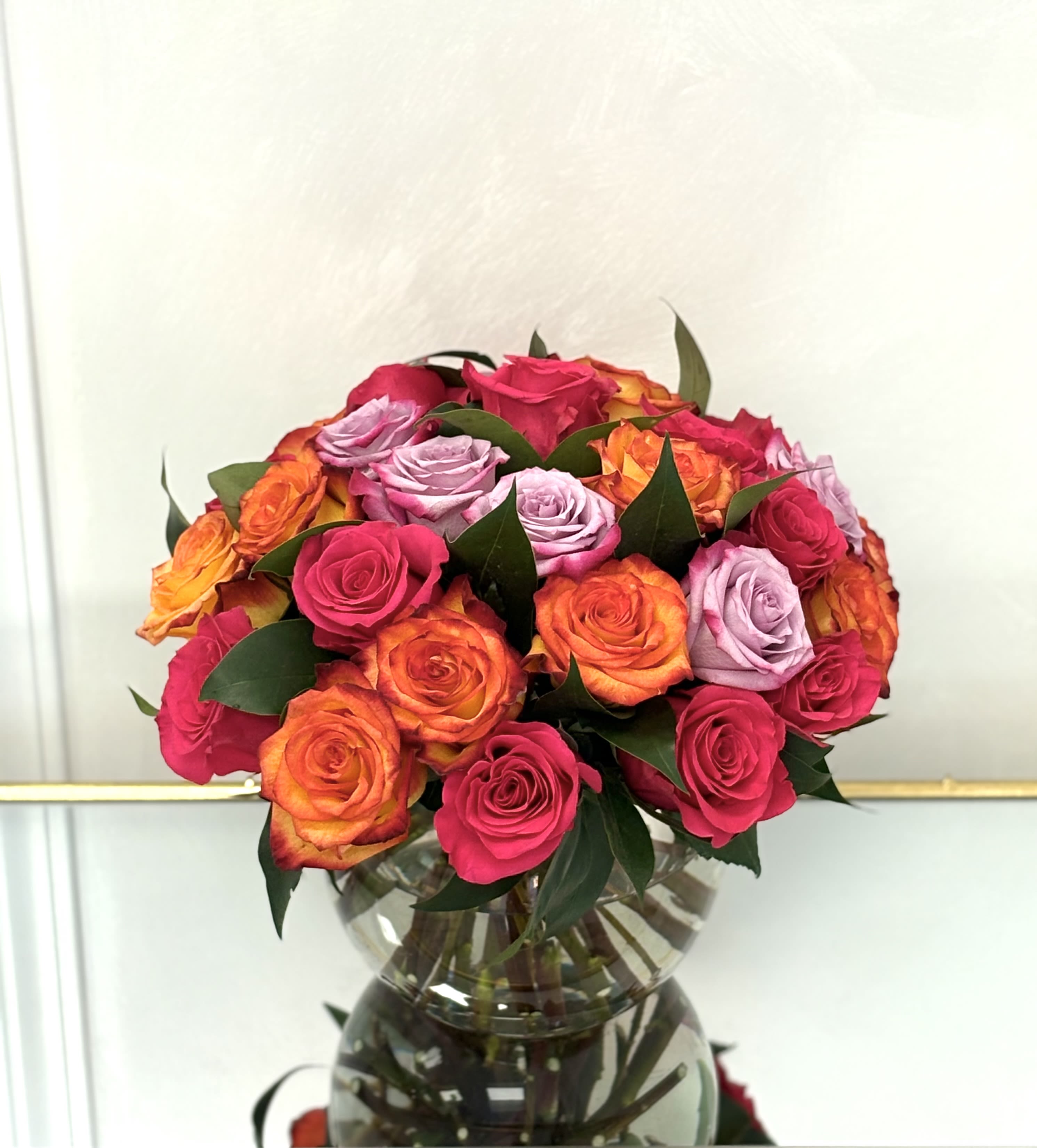 Round bouquet of hot pink, orange, and lavender roses arranged in a clear glass vase.