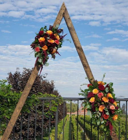 Autumn-toned floral sprays of red and yellow roses on a rustic wooden triangle frame outdoors.