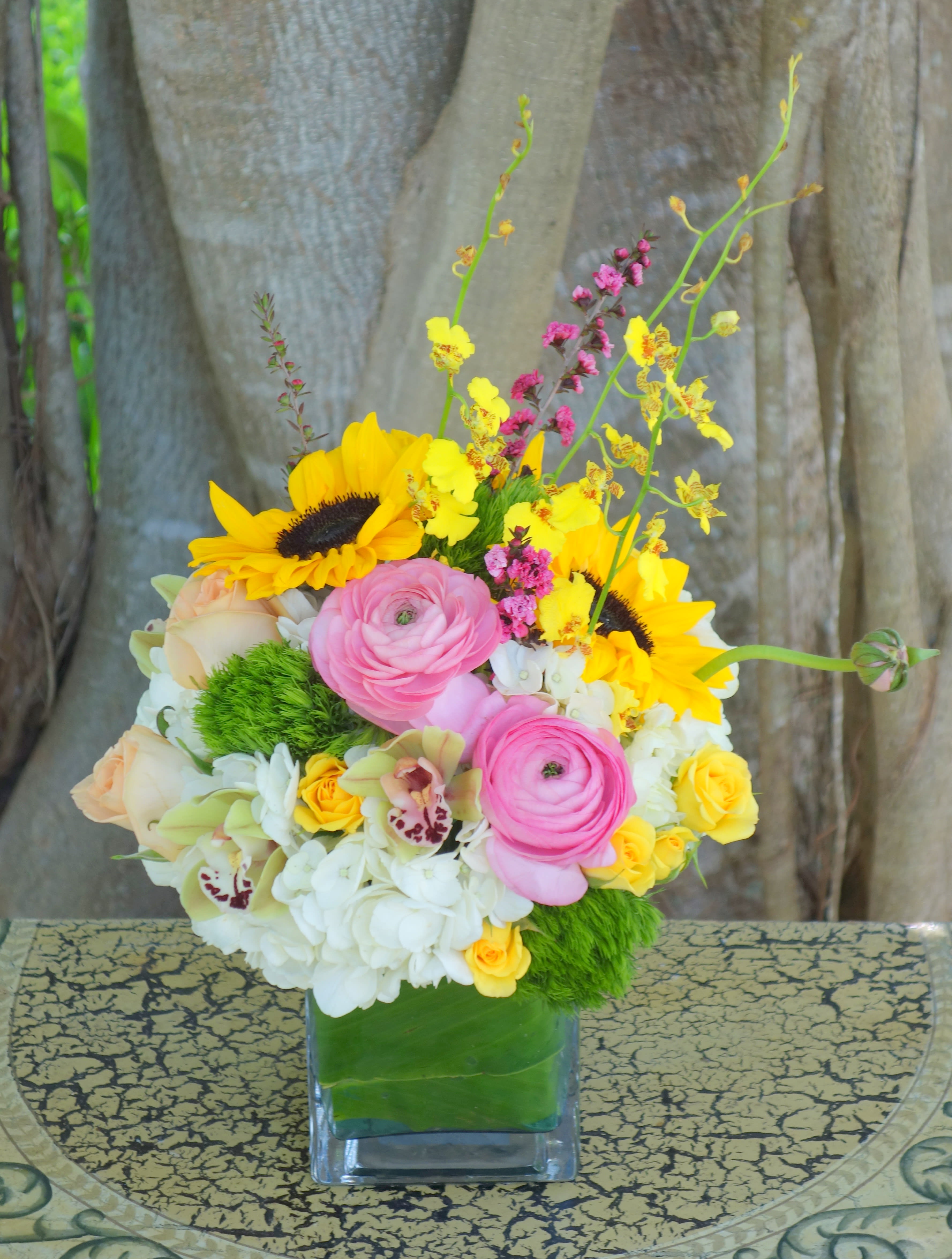 Bright mixed bouquet with sunflowers, pink ranunculus, orchids, and hydrangeas in a glass cube vase