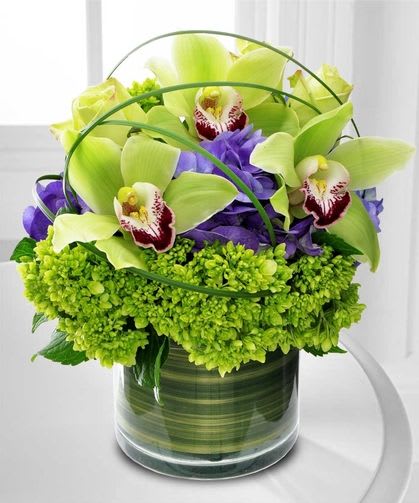 Compact arrangement of green cymbidium orchids and purple flowers in a clear glass cylinder vase