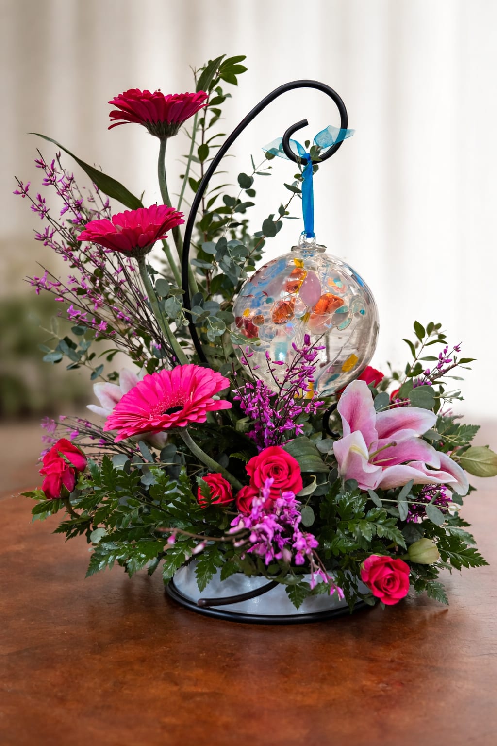 Glass Orb with Designer's Choice Flowers - Please Note: Flowers are designer’s choice and will vary based on seasonal availability to ensure the freshest and most beautiful selection. If you have specific flower or color requests, please call us at (574) 722-1155 and we’ll be happy to customize your order.  Add a meaningful touch of beauty to any space with our handcrafted 6-inch glass ornament display, thoughtfully designed with a fresh floral arrangement. Each piece features a stunning hand-blown glass orb suspended on an elegant stand, surrounded by a lush mix of seasonal blooms and greenery.  Perfect as a heartfelt gift, memorial piece, or decorative accent, this arrangement brings together artistry and nature in a unique and lasting way. The vibrant glass ornament catches the light beautifully, while the fresh flowers add softness and warmth.  Details: 6” hand-blown glass ornament on a decorative stand Fresh floral arrangement included Approximate overall size may vary based on design Ideal for gifts, sympathy, or home décor 